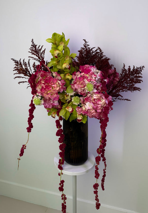 Floral arrangement with pink and green flowers in a black vase on a white pedestal against a white wall.