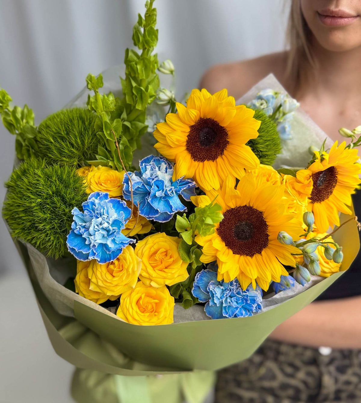 A vibrant flower bouquet featuring yellow sunflowers, yellow roses, blue carnations, and green filler flowers, wrapped in light green paper and held by a person whose face and arm are partially visible in a softly lit indoor setting.