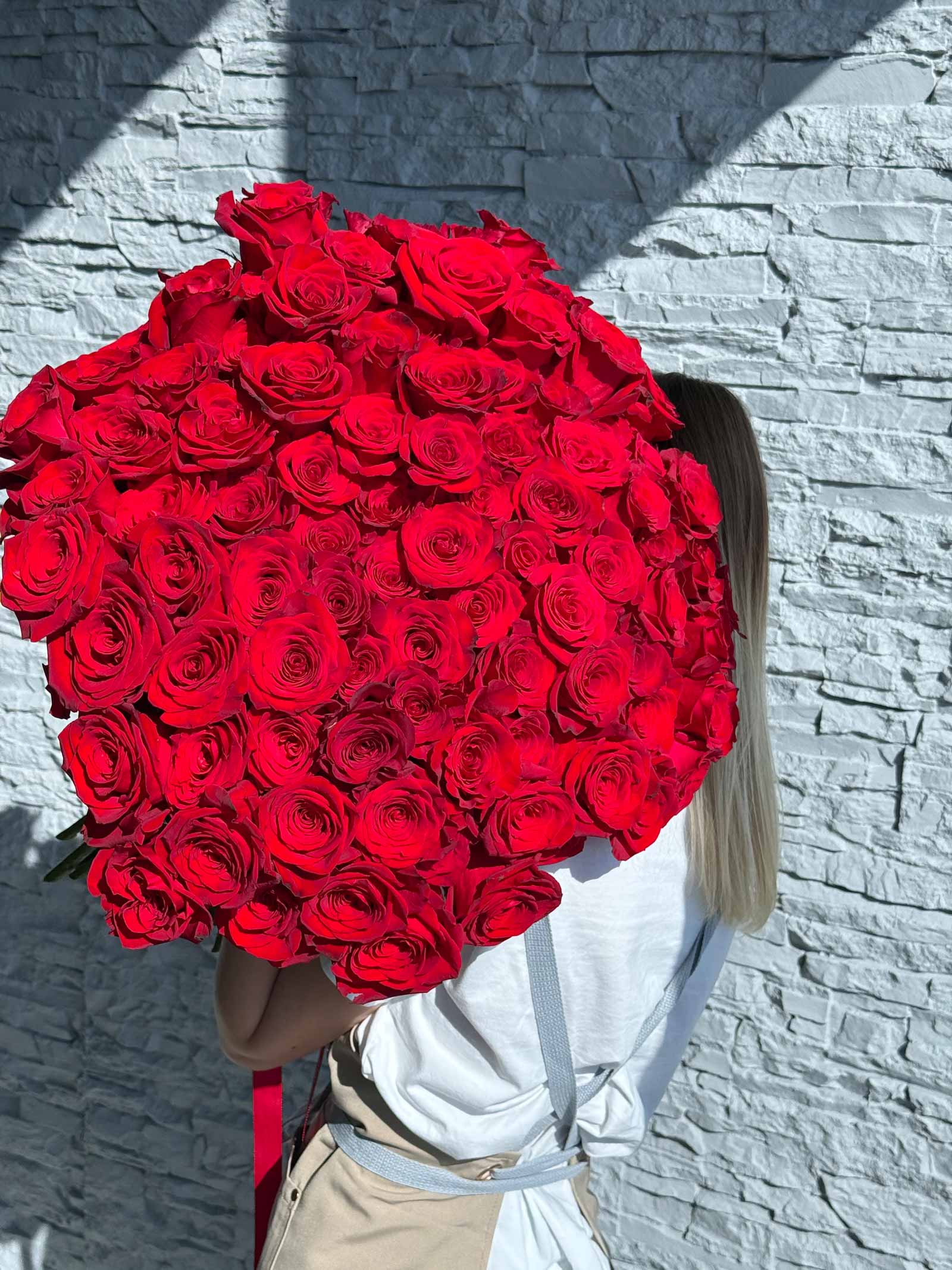 Woman holding a large bouquet of red roses in front of a white brick wall in bright sunlight.