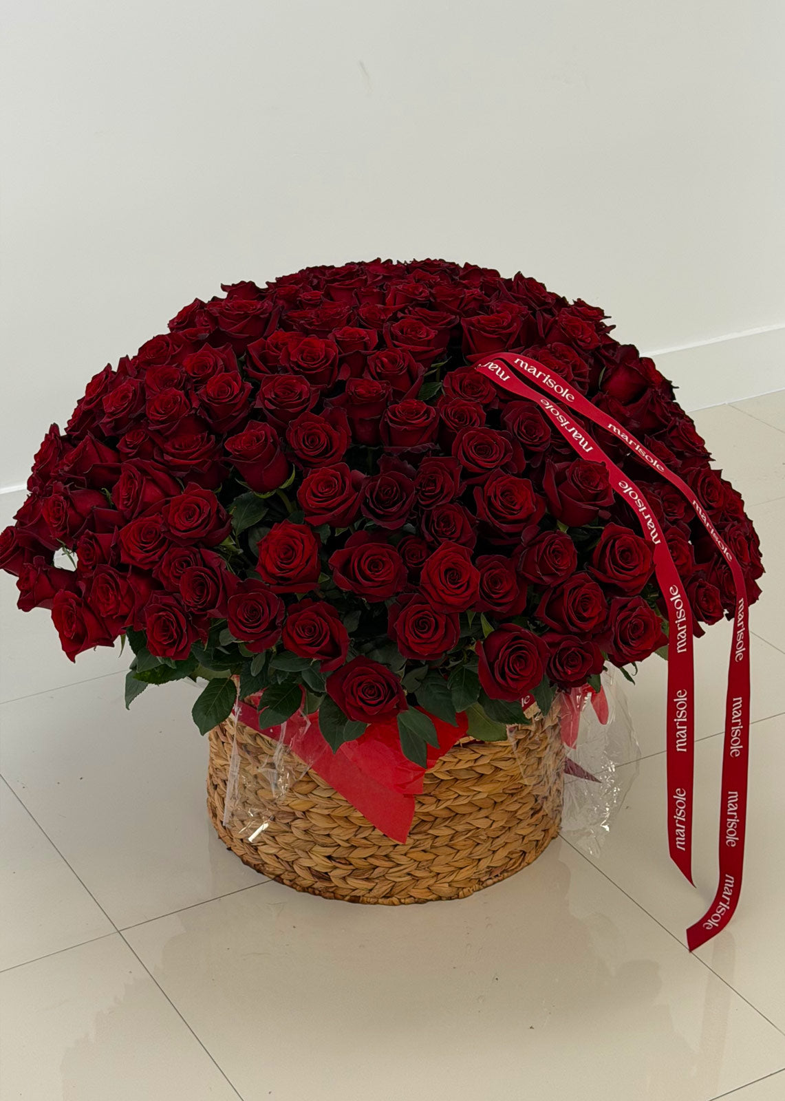 Bouquet of red roses in a woven basket with a red ribbon on a white floor.