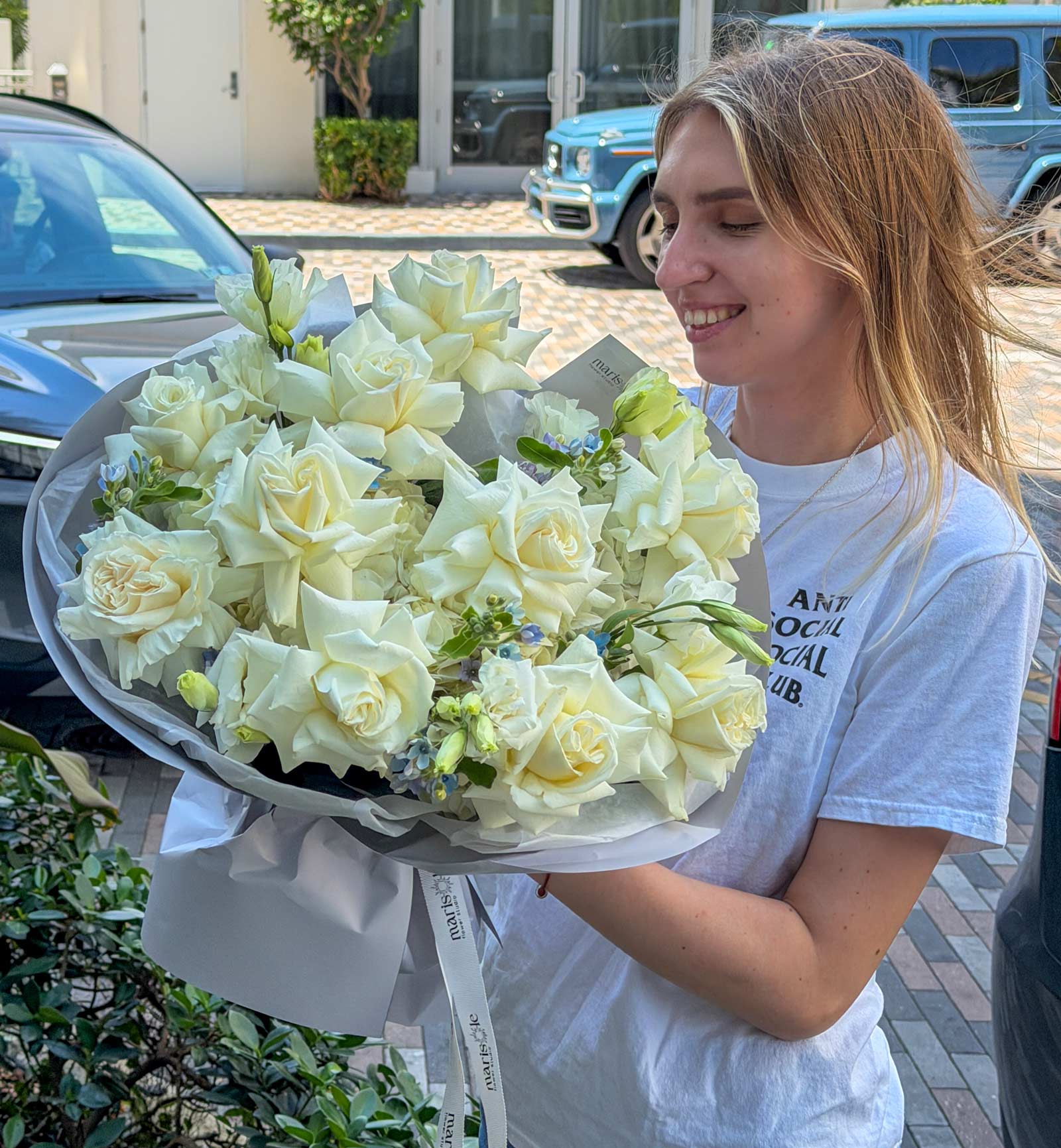 Woman holding a bouquet of white flowers outdoors with cars and a building in the background.