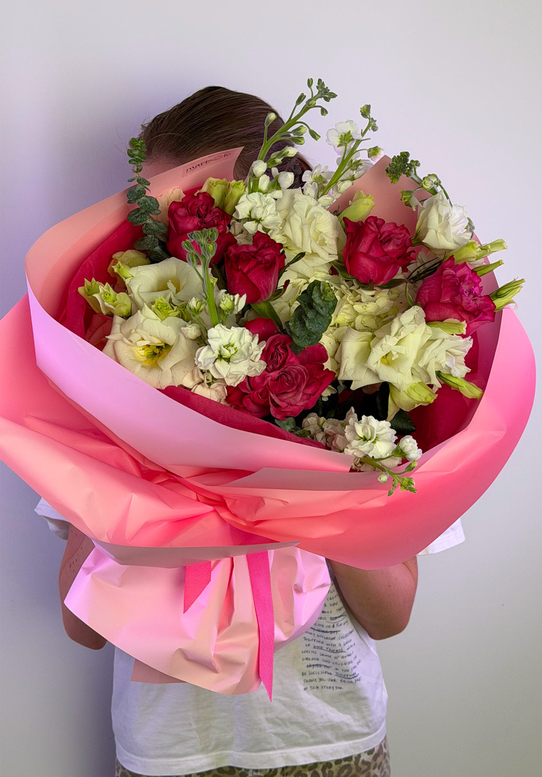 Person holding a bouquet of flowers wrapped in pink paper against a plain background