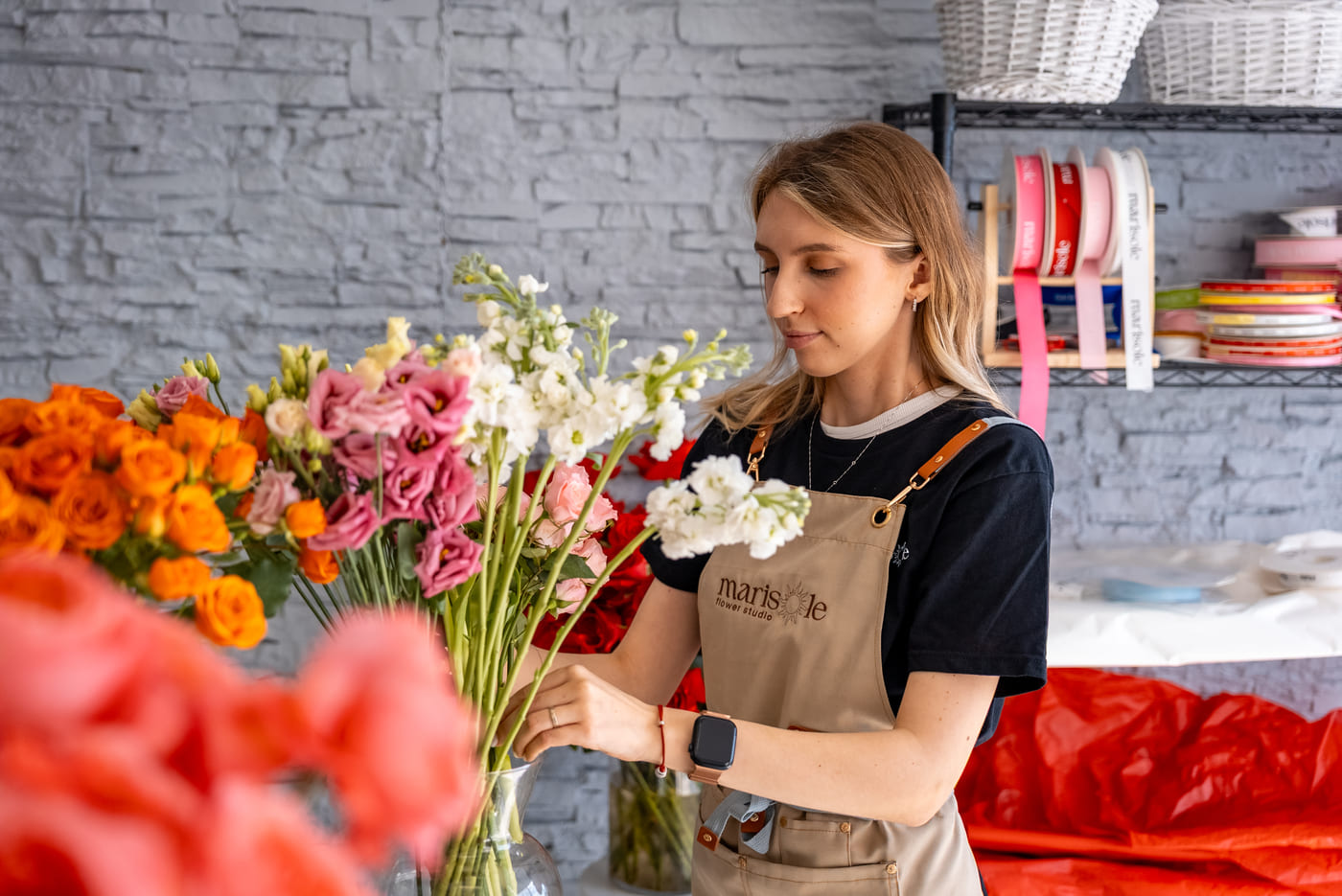 Woman arranging flowers in a floral shop with a gray brick wall background.