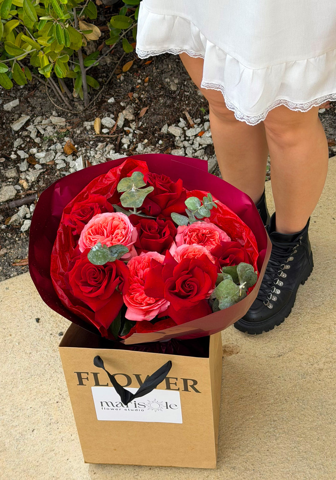 Bouquet of red roses in a cardboard box with 'Marisole Flowers' branding, held by a person wearing a white dress and black boots.