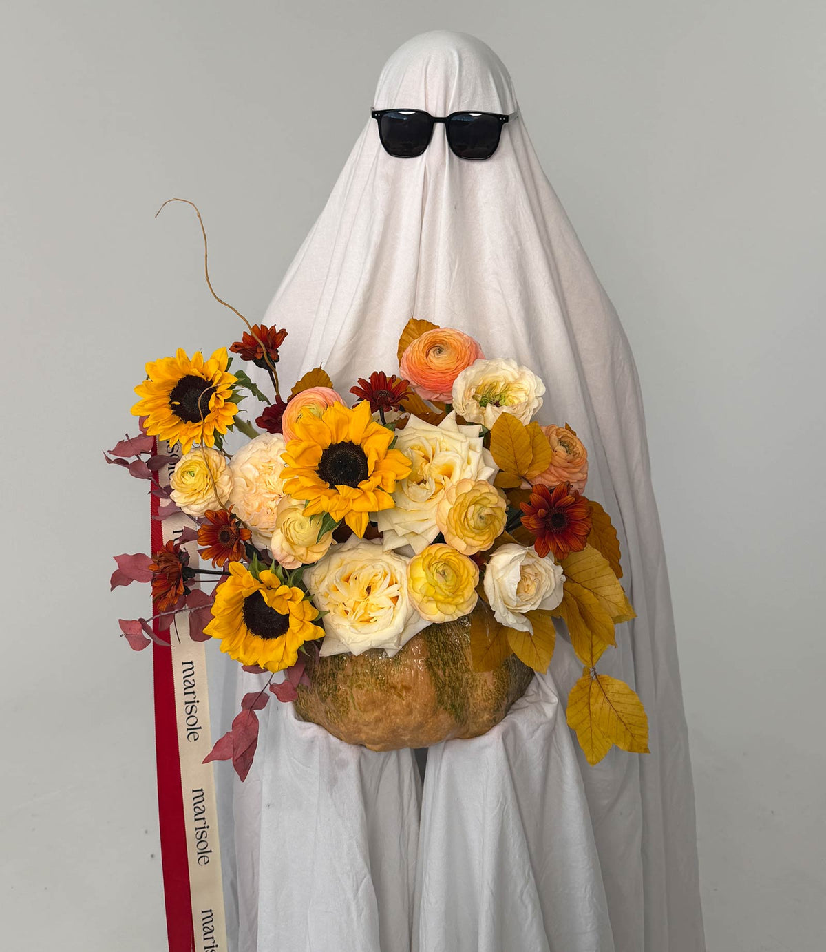 A ghostly Halloween decoration or costume prop made from a sheet-covered form wearing black sunglasses and holding a large autumn flower arrangement in a pumpkin-shaped vase, photographed against a plain light gray background.