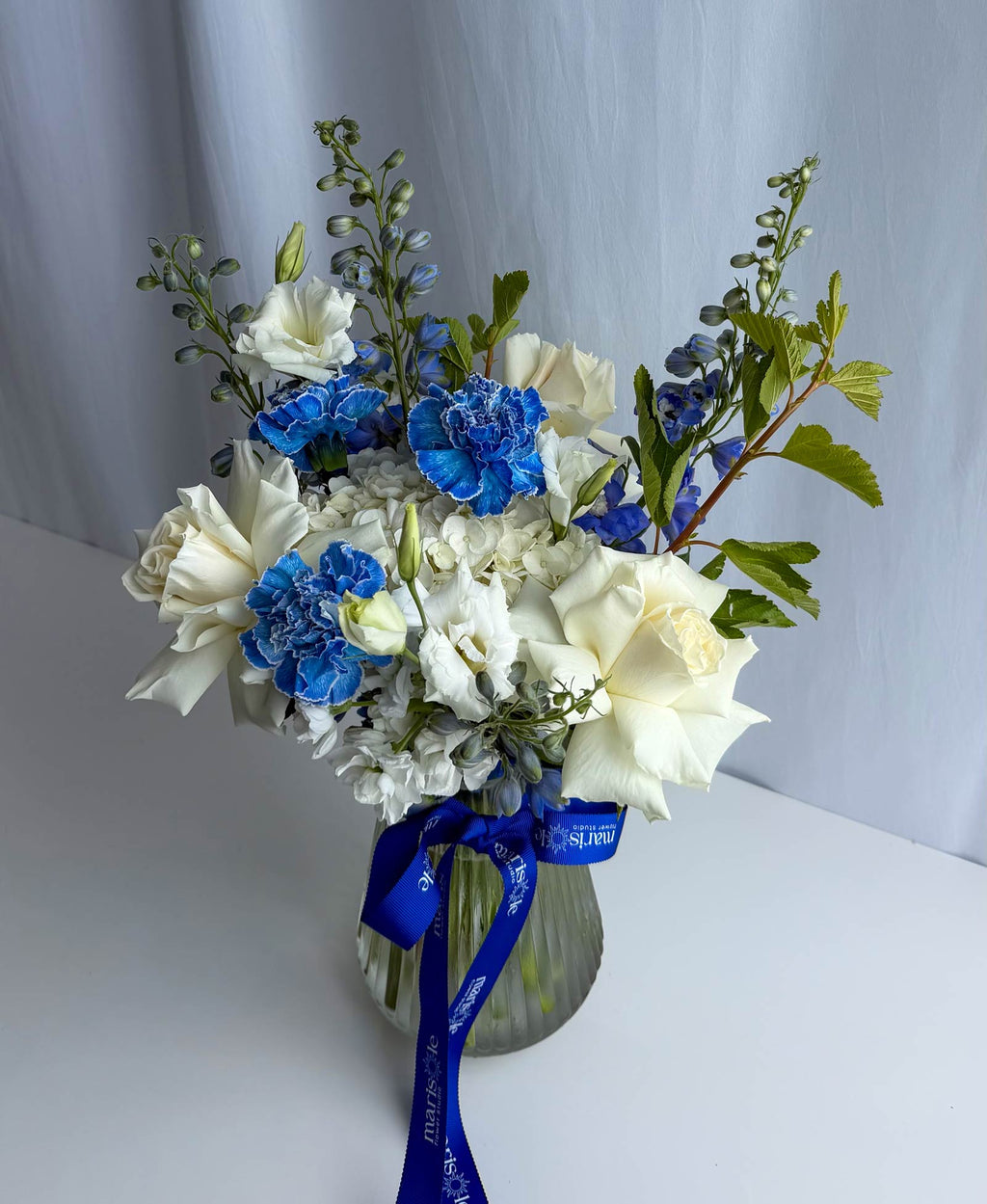 A floral arrangement in a ribbed glass vase featuring white roses, blue carnations, blue delphiniums, and green foliage, tied with a blue ribbon