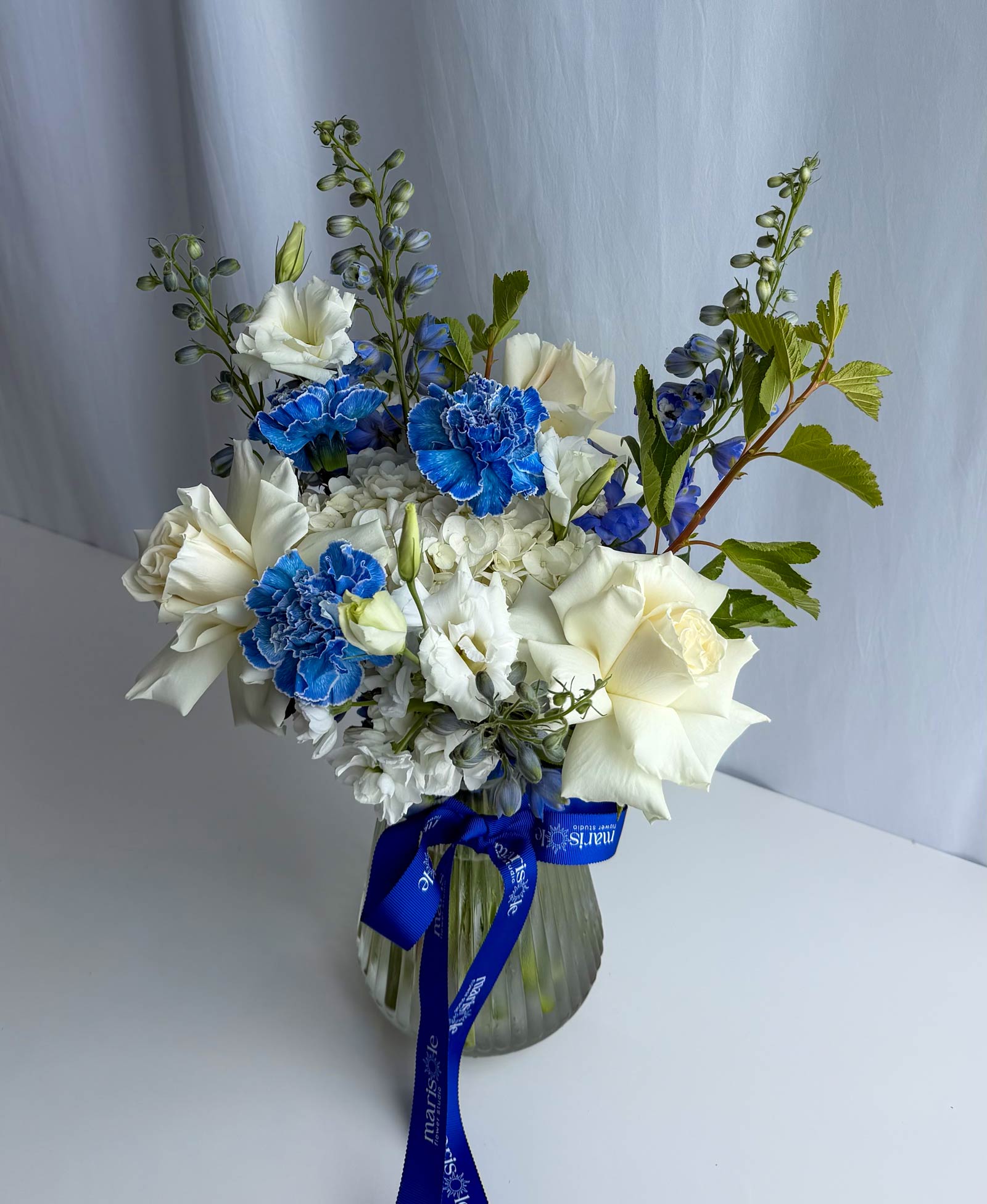 A floral arrangement in a ribbed glass vase featuring white roses, blue carnations, blue delphiniums, and green foliage, tied with a blue ribbon
