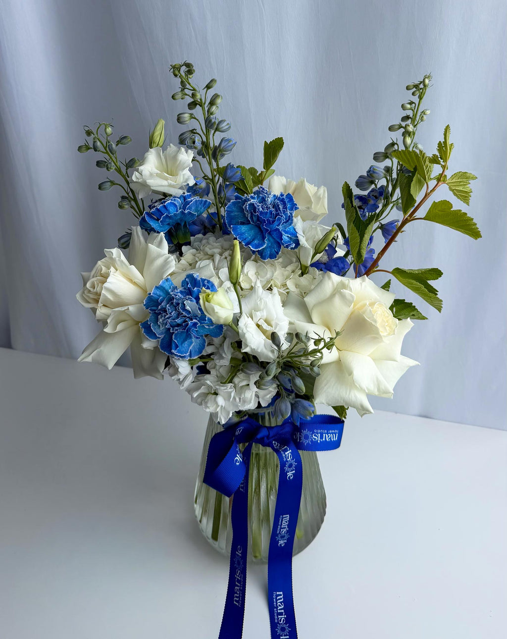 A floral arrangement in a clear glass vase with a blue ribbon, featuring white roses, blue carnations, white lisianthus, and green foliage, set against a light gray draped backdrop.