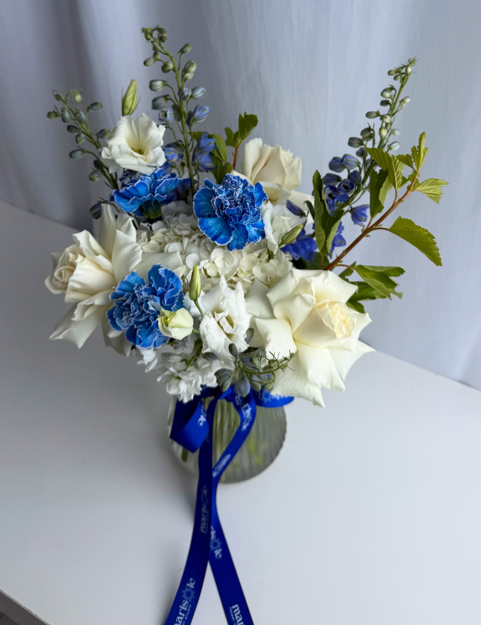 A floral arrangement in a clear glass vase featuring white roses, blue carnations, white hydrangeas, and green foliage, tied with a blue ribbon on a white surface against a light gray backdrop.