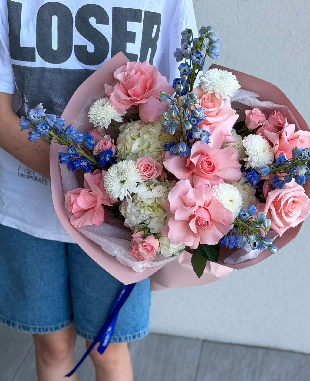 Blush & Sky Bouquet with hydrangea, French roses, delphinium, spray roses, and chrysanthemums in premium wrapping.