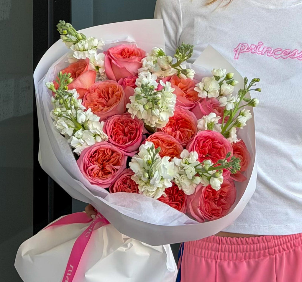 A close-up of a bouquet of pink and coral roses with white filler flowers wrapped in white paper, held by a person wearing a white T-shirt with pink lettering and pink shorts.