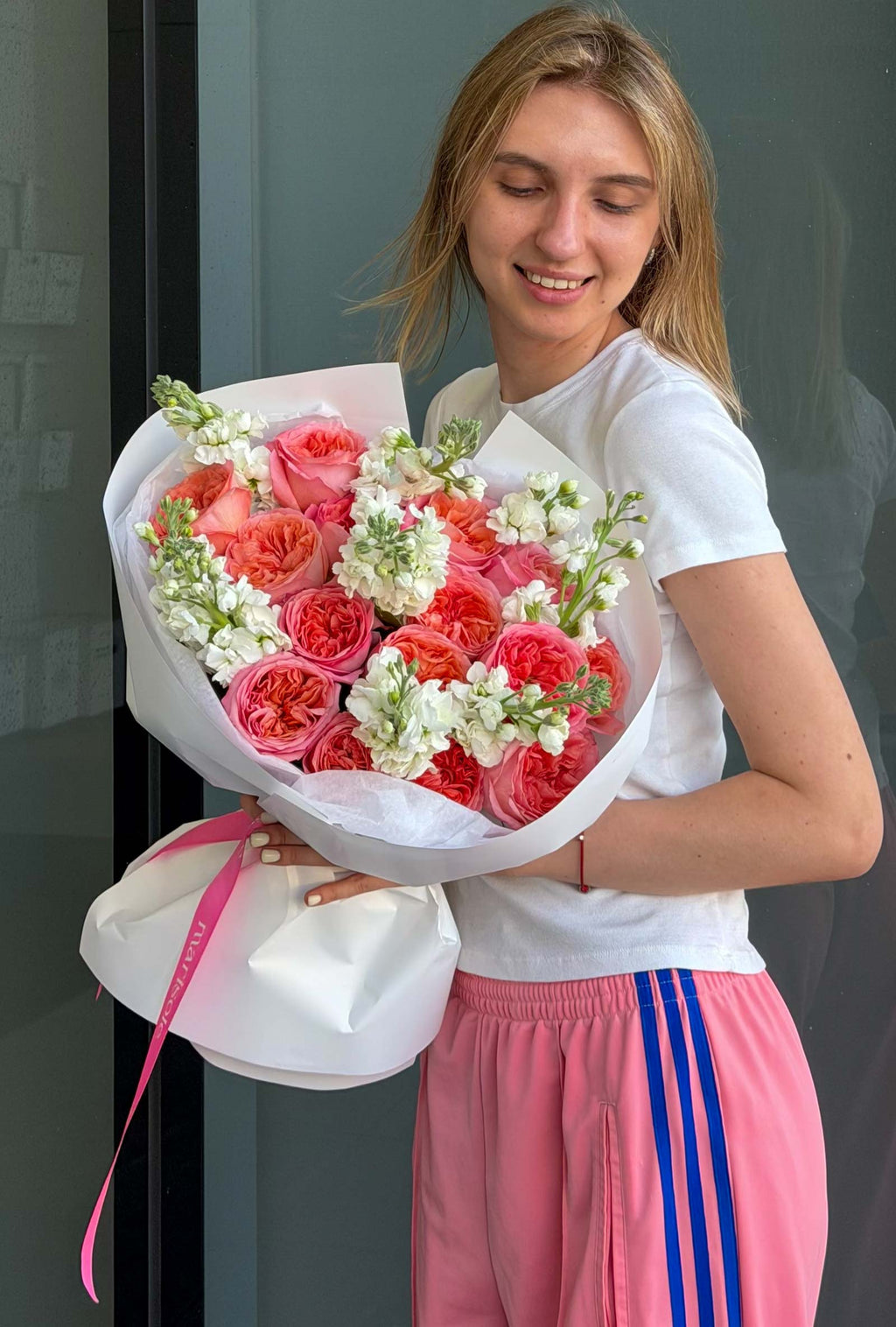 A smiling young woman with blonde hair holds a large bouquet of pink roses with white filler flowers. 