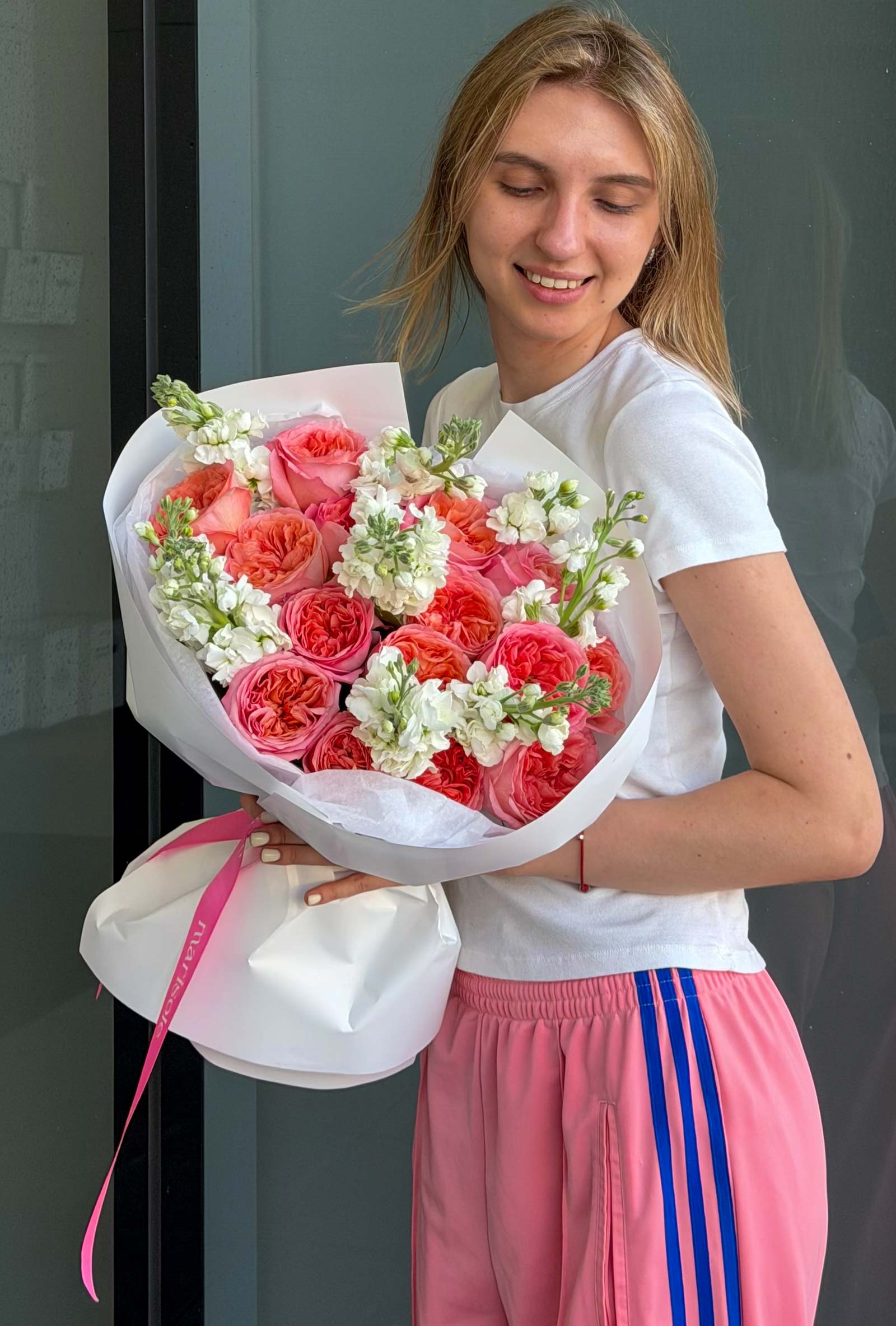 A smiling young woman with blonde hair holds a large bouquet of pink roses with white filler flowers. 