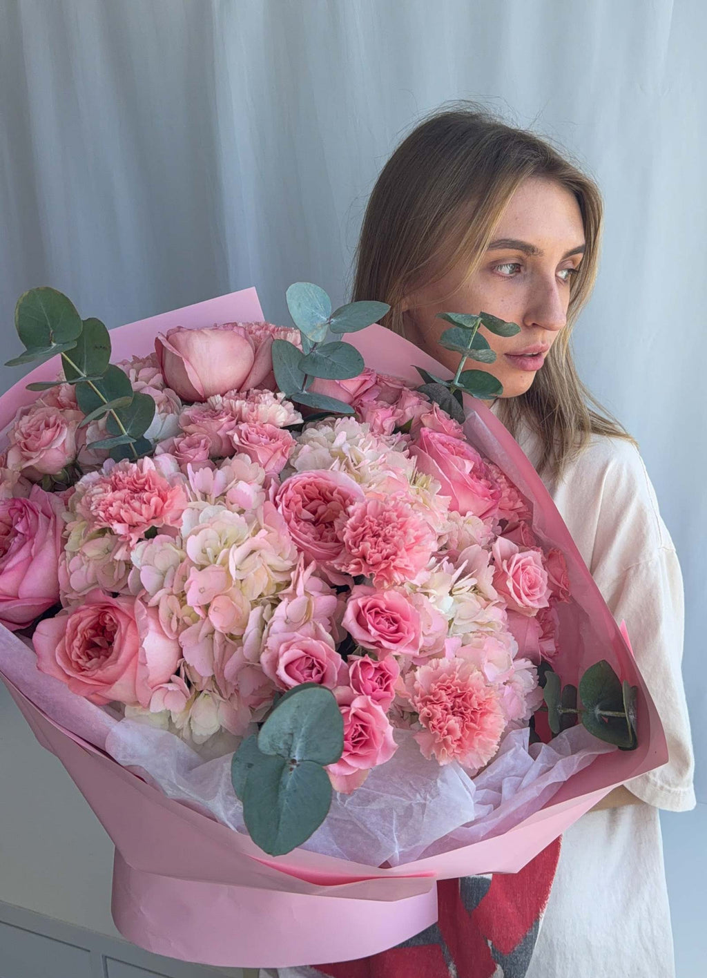 A woman holding a large, vibrant bouquet of pink and white flowers with green eucalyptus leaves, wrapped in pink paper, against a soft light gray curtain background.