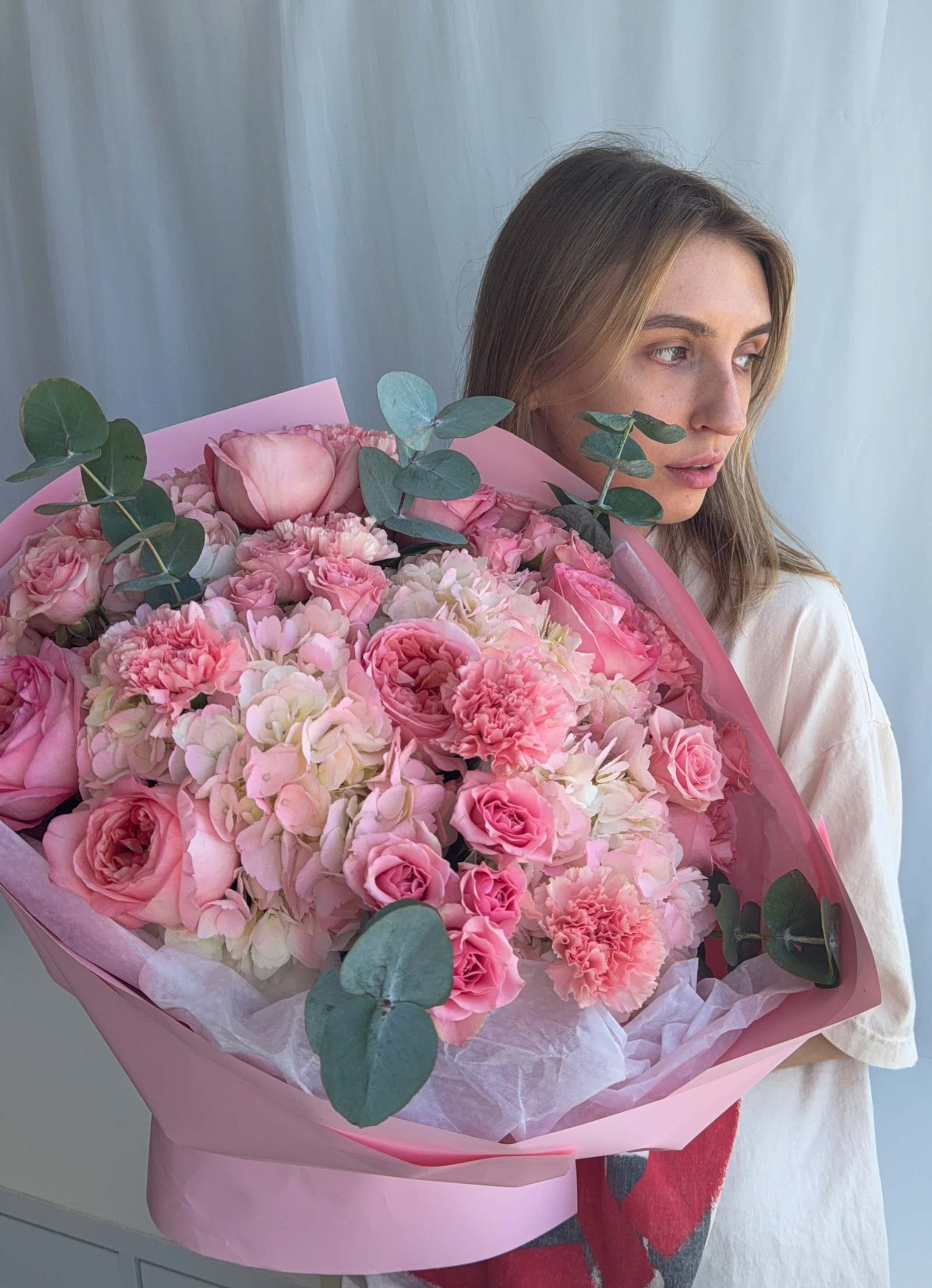 A woman holding a large, vibrant bouquet of pink and white flowers with green eucalyptus leaves, wrapped in pink paper, against a soft light gray curtain background.