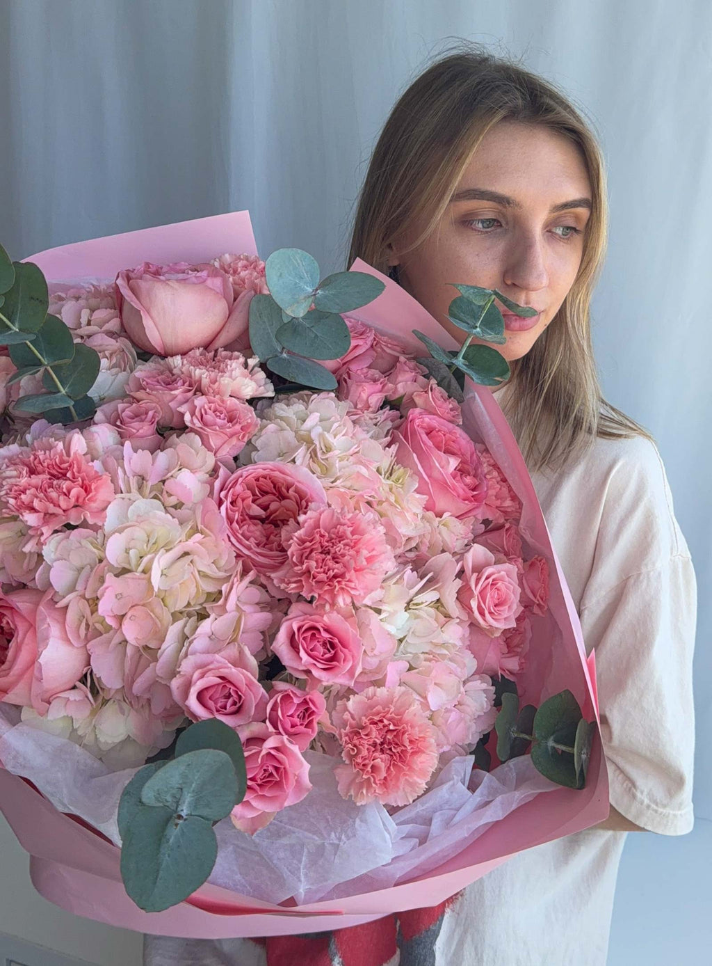A woman holding a large, vibrant bouquet of pink flowers, including roses, carnations, and hydrangeas, wrapped in pink paper with green eucalyptus leaves, against a light gray curtain background.
