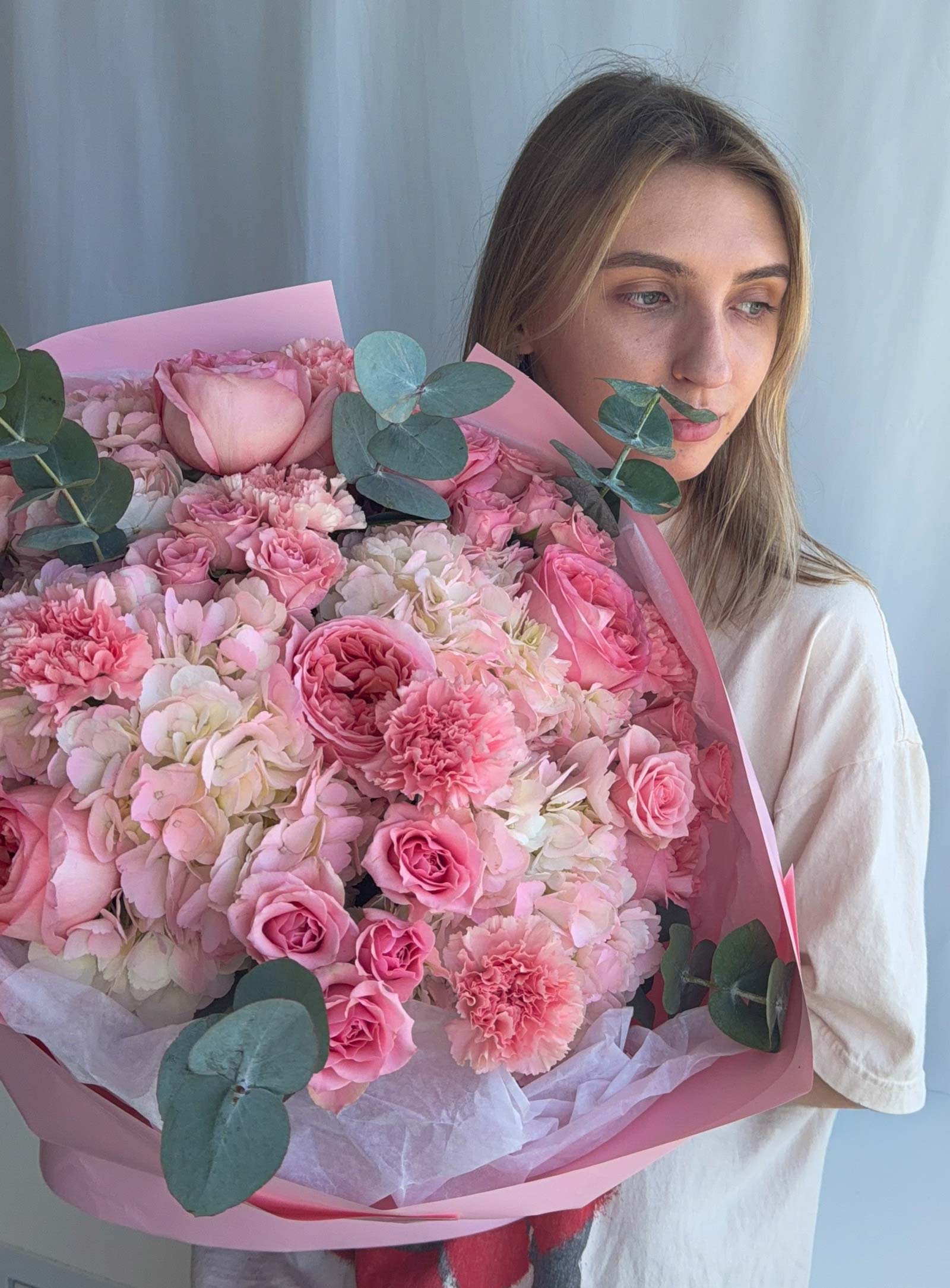 A woman holding a large, vibrant bouquet of pink flowers, including roses, carnations, and hydrangeas, wrapped in pink paper with green eucalyptus leaves, against a light gray curtain background.