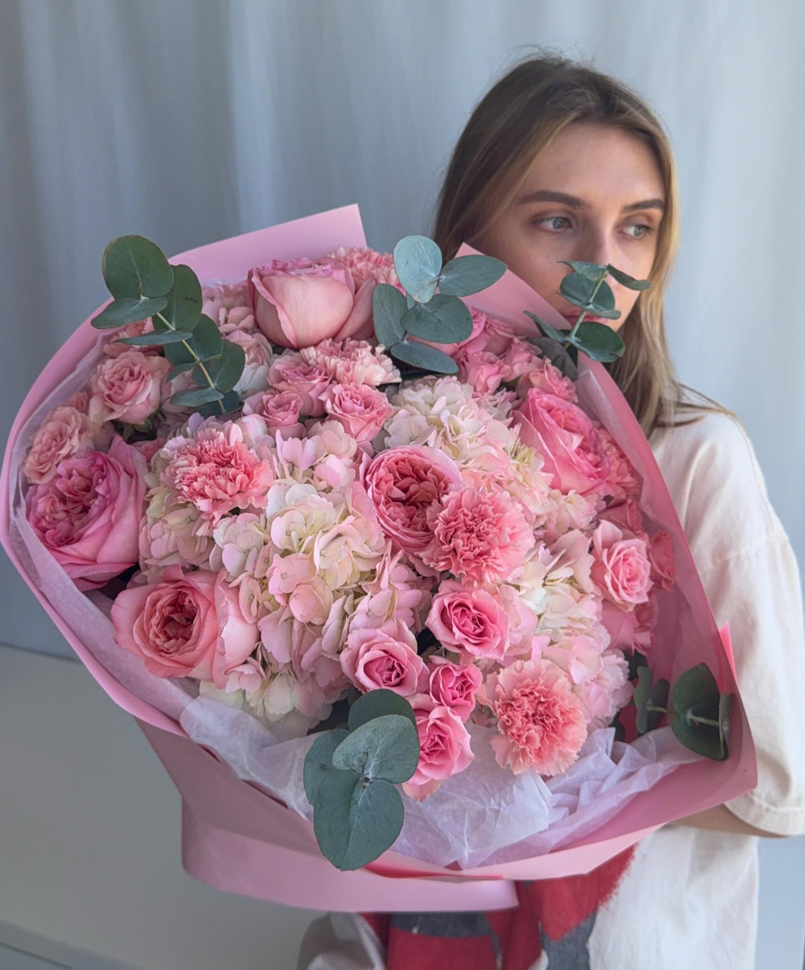 A large, vibrant bouquet of pink and light pink flowers, including roses and hydrangeas, wrapped in pink tissue paper with green eucalyptus leaves, held by a woman in a white top against a light gray curtain background.