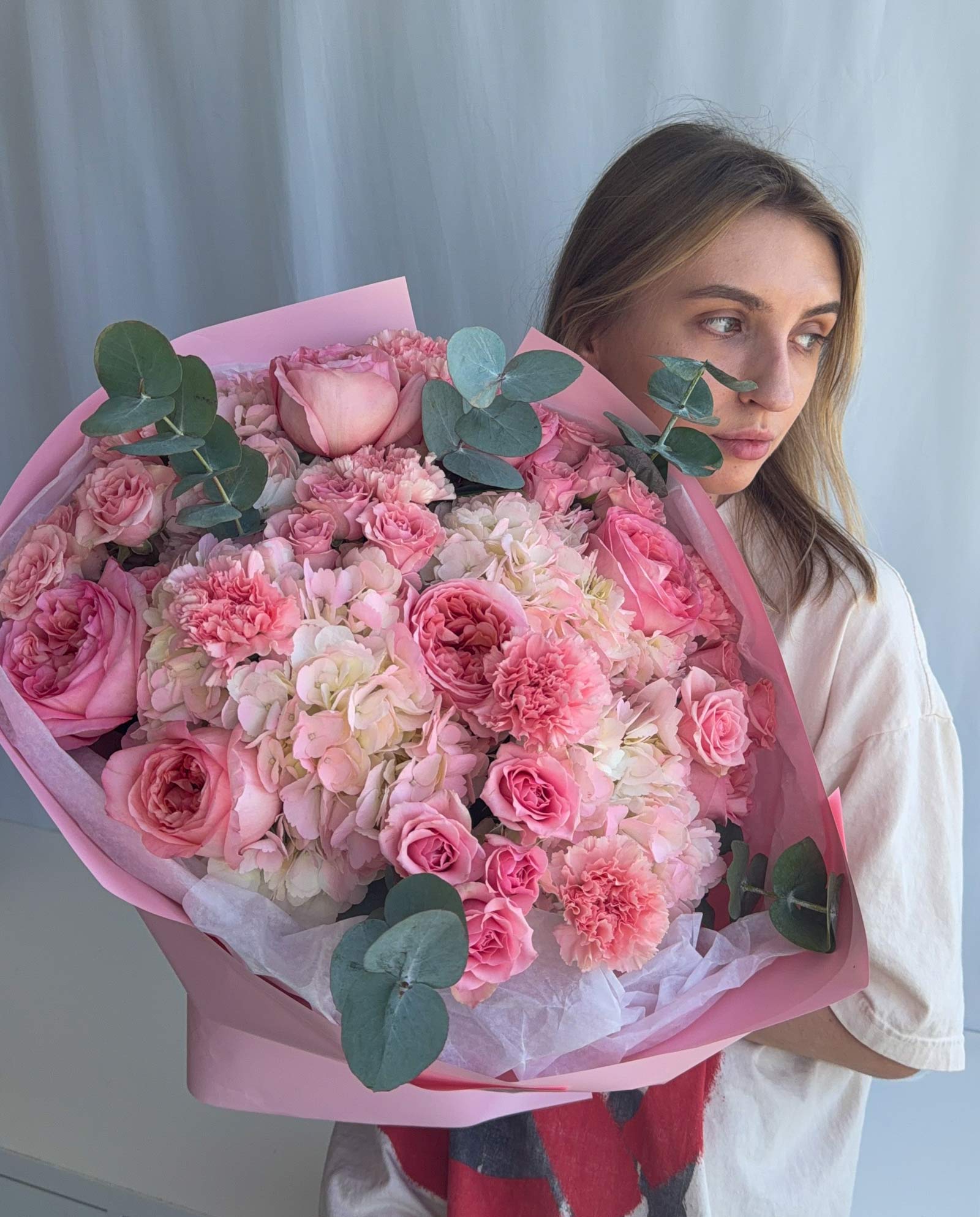 A woman holding a large, vibrant pink flower bouquet with pink roses, carnations, and green eucalyptus leaves, wearing a white shirt against a light gray curtain background.