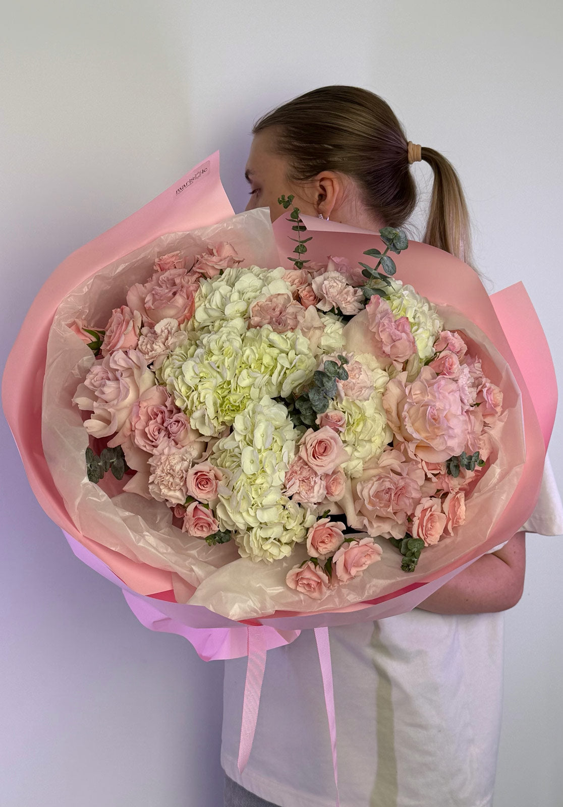 Person holding a large bouquet of pink and white flowers wrapped in pink paper against a plain background