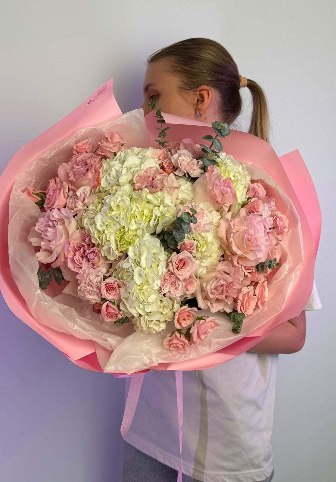 Person holding a large bouquet of pink and white flowers wrapped in pink paper against a plain background