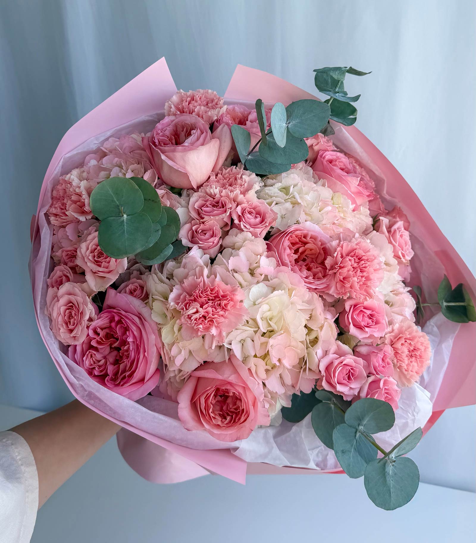 A large, beautifully arranged bouquet of pink and white flowers, including roses, carnations, and hydrangeas, wrapped in pink tissue paper and held by a person's hand against a soft blue curtain background.