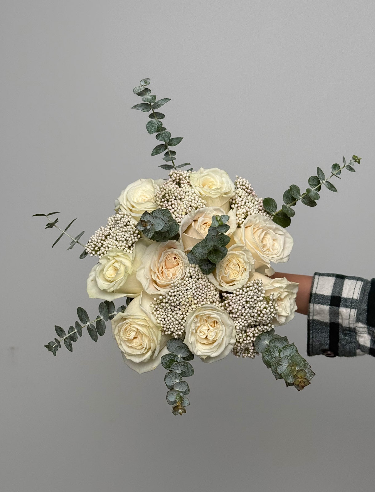 Bouquet of white roses with greenery held by a person wearing a plaid shirt against a gray background