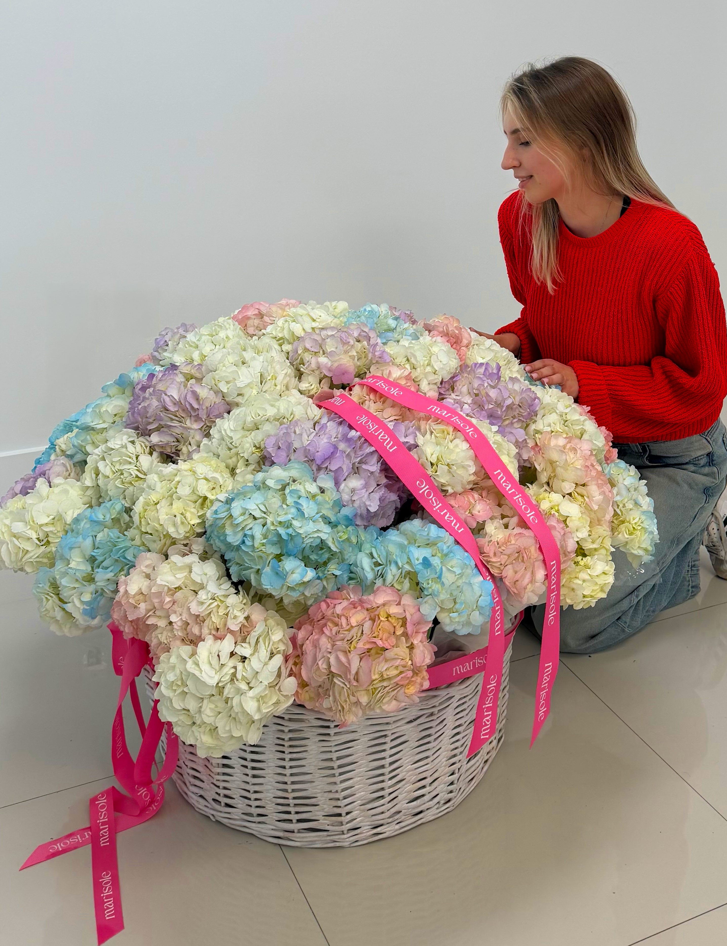 Woman holding a large bouquet of pastel-colored flowers in a wicker basket with pink ribbons.