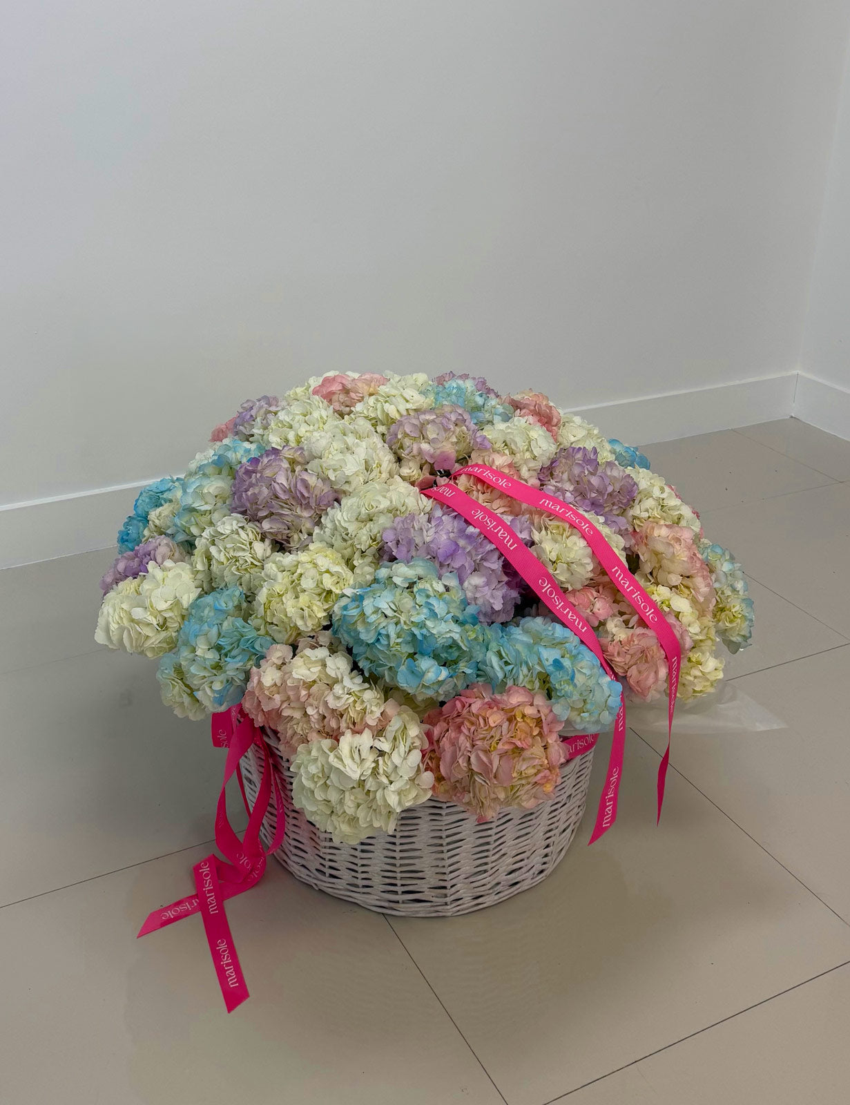 Colorful bouquet of flowers in a white basket with pink ribbons on a light-colored floor.