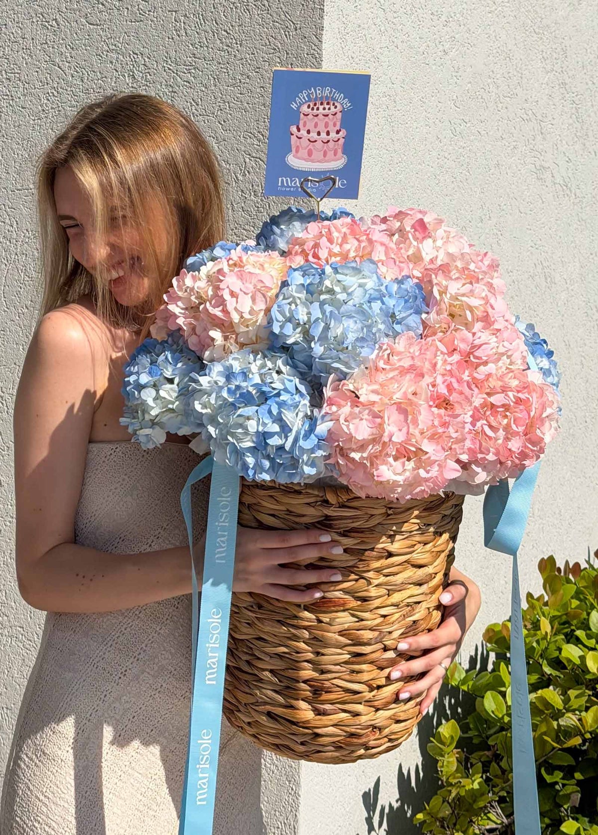 A woman in a beige dress holds a large woven basket filled with pastel pink, blue, and light blue hydrangeas.