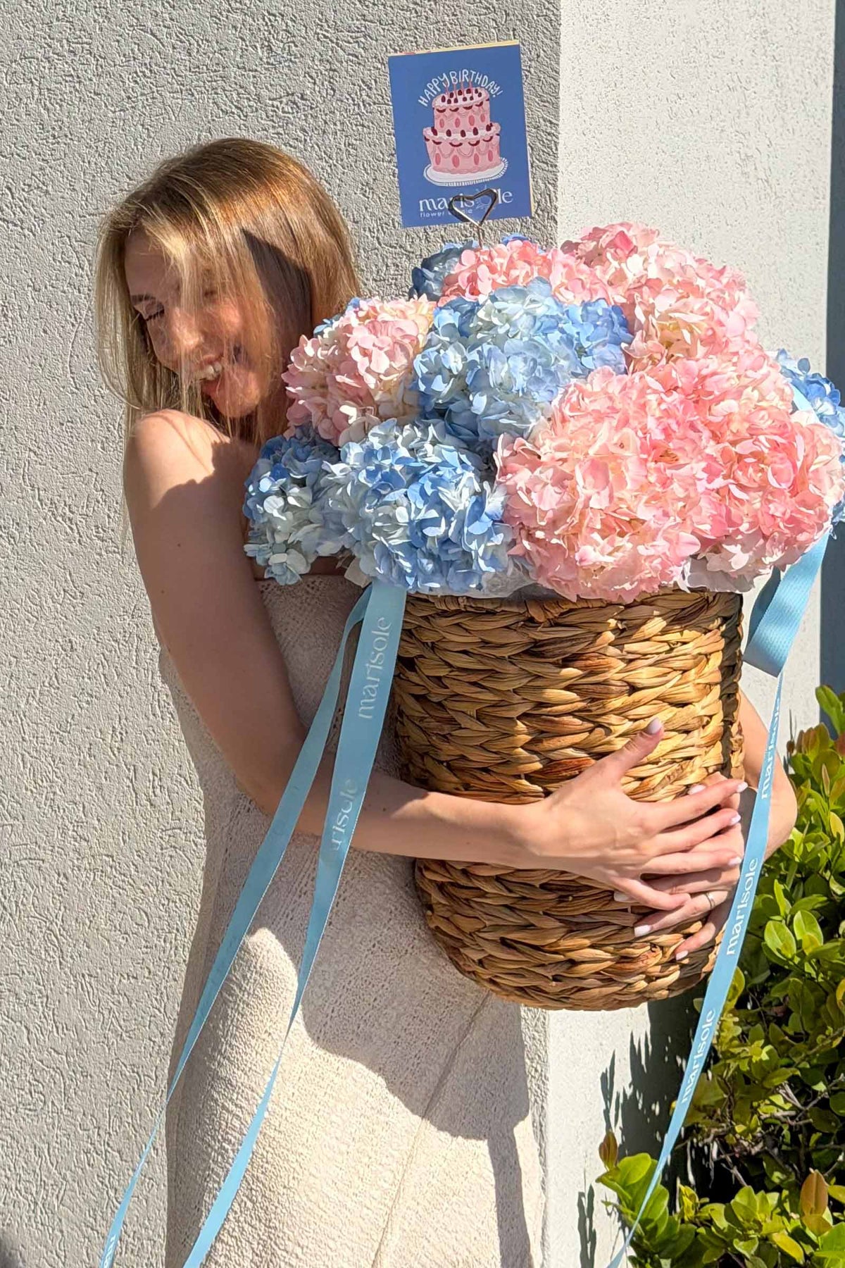 A woman in a beige dress holds a large woven basket filled with pink and blue hydrangea blooms tied with light blue ribbons.