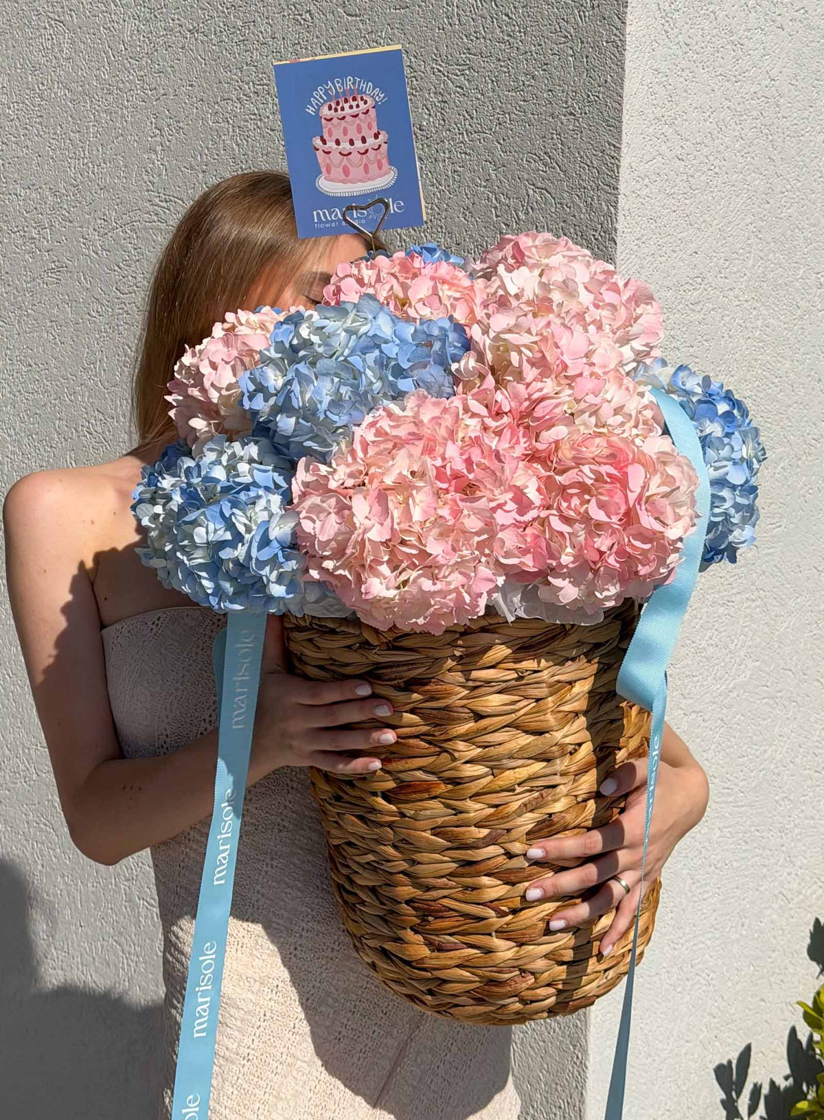 A person holding a large woven basket filled with pink and blue hydrangea blooms, with a blue ribbon and a small birthday card attached.