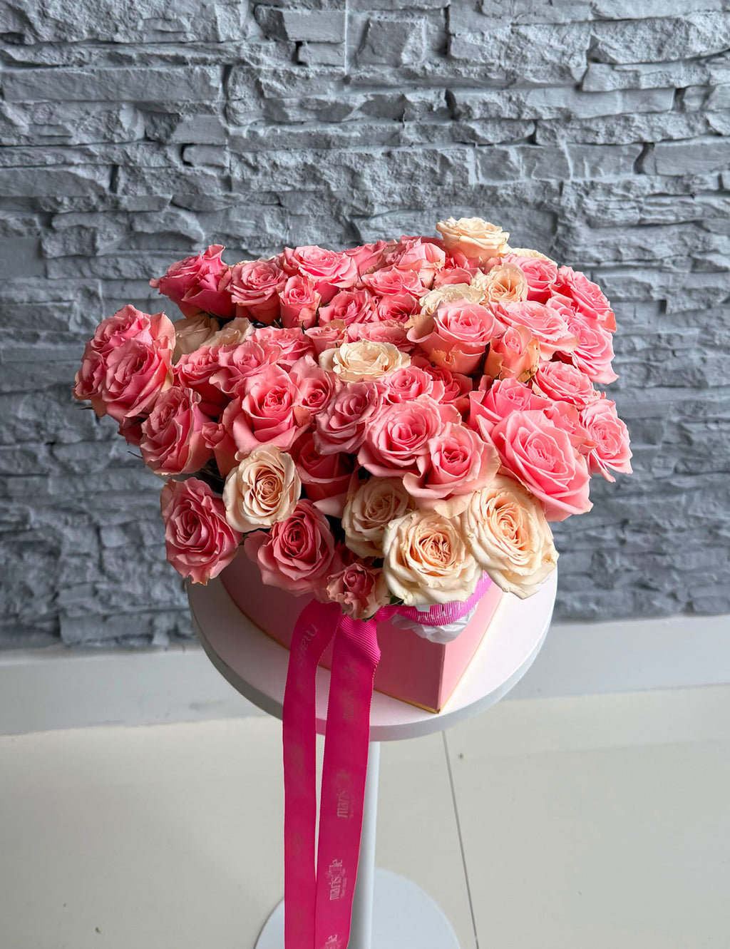 Bouquet of pink and beige roses in a box with a pink ribbon against a gray stone wall.
