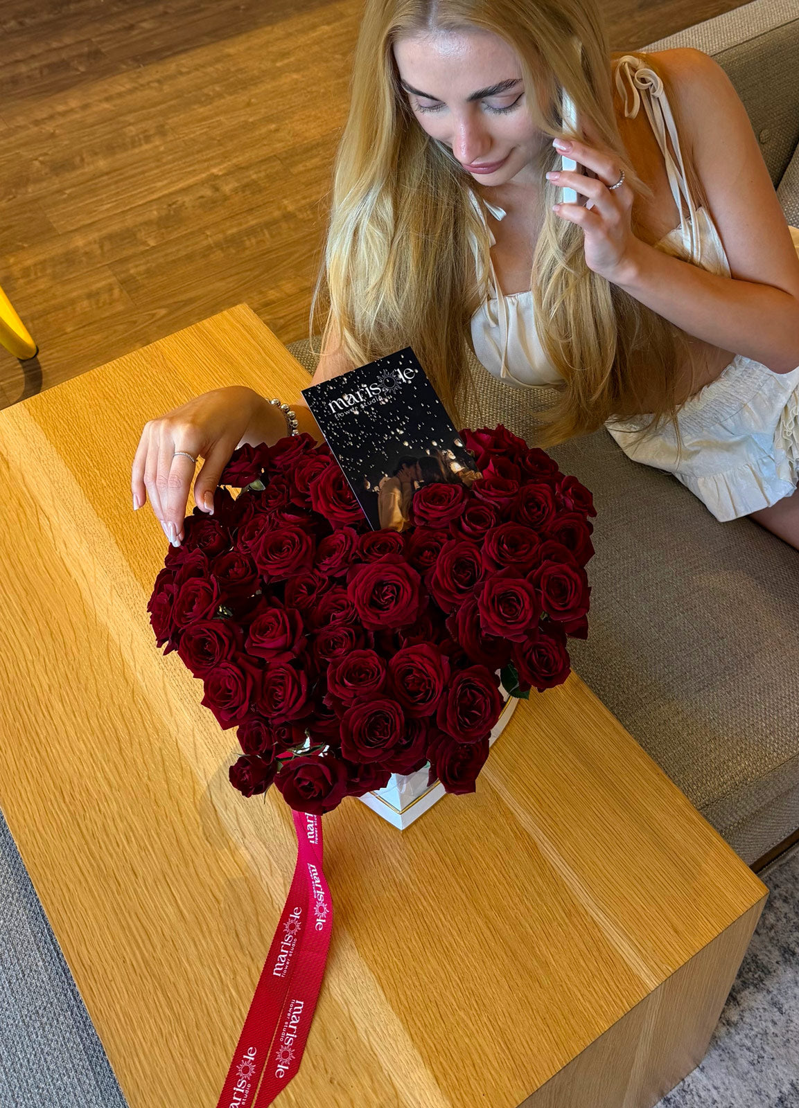 Woman sitting on a couch with a large bouquet of red roses and a black box on a wooden table.