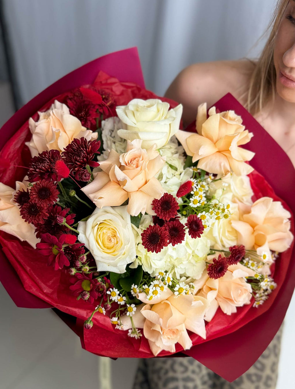 A large, vibrant bouquet of mixed flowers wrapped in deep red tissue paper, held by a person whose face and shoulder are partially visible in the background.