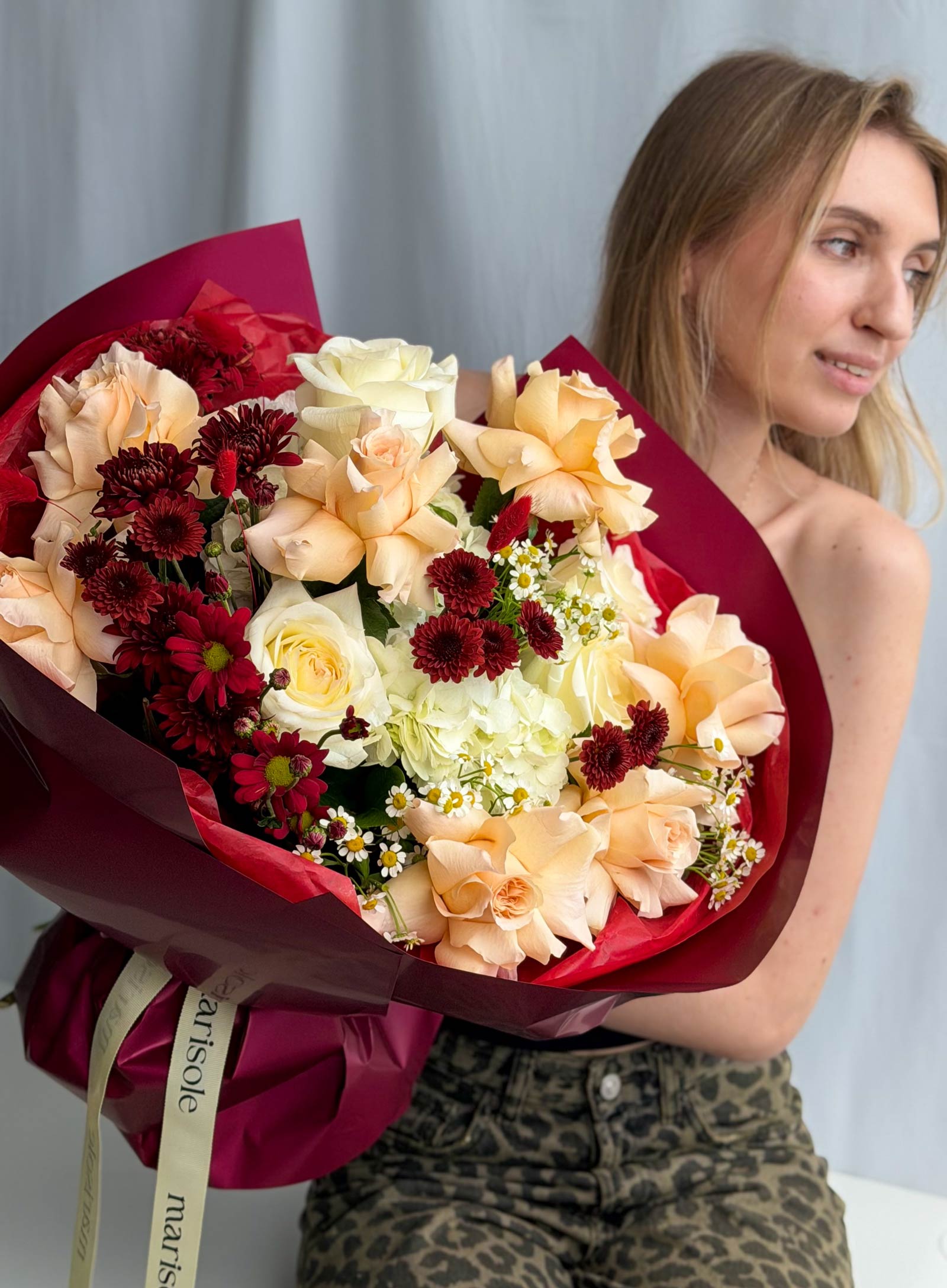 A woman holding a large, vibrant bouquet of mixed flowers, including peach roses, cream roses, deep red chrysanthemums, and white daisies, wrapped in deep red paper with a cream ribbon.
