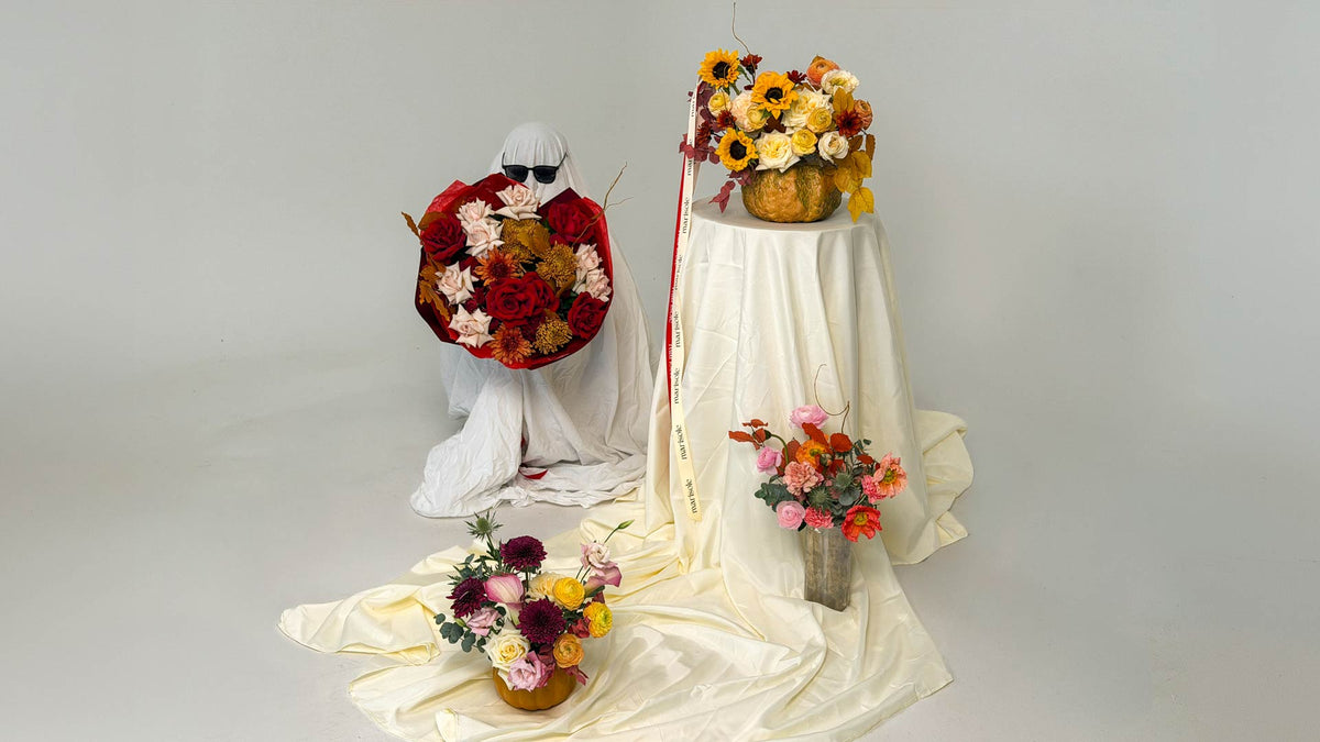 A styled floral arrangement display on a light gray studio background, featuring a white draped pedestal with a bouquet of yellow, orange, and red flowers, a kneeling figure in white holding a red and cream floral arrangement, and smaller flower arrangements on draped cream fabric.