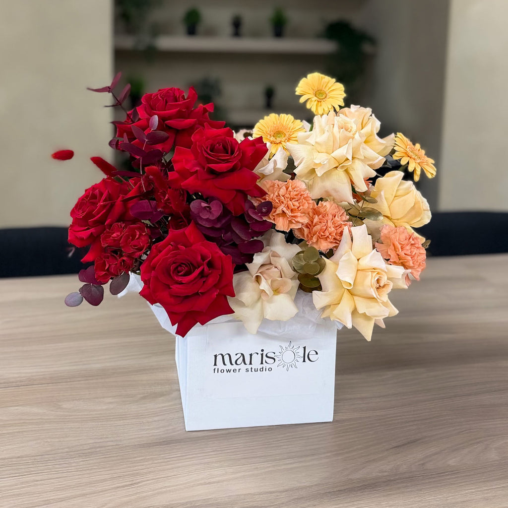 A floral arrangement in a white square box labeled 'marisole flower studio,' featuring deep red roses, cream and peach flowers, yellow daisies, and green foliage, displayed on a light wood tabletop.