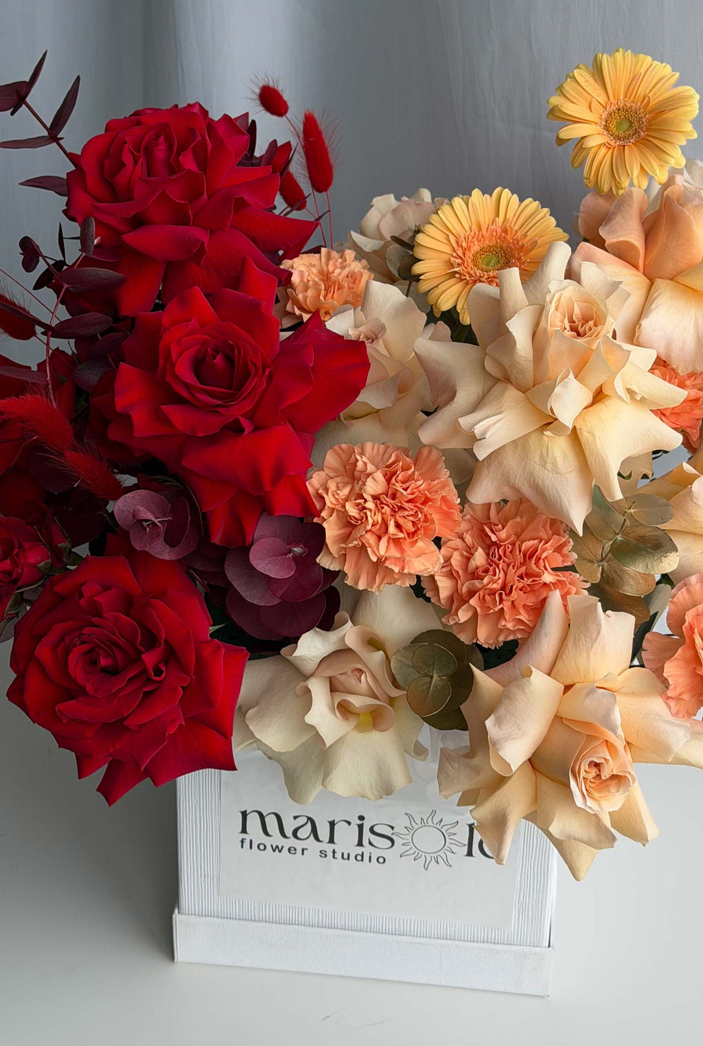 A vibrant floral arrangement featuring deep red roses, peach carnations, cream roses, and yellow gerbera daisies in a white wooden box labeled 'marisole flower studio'.