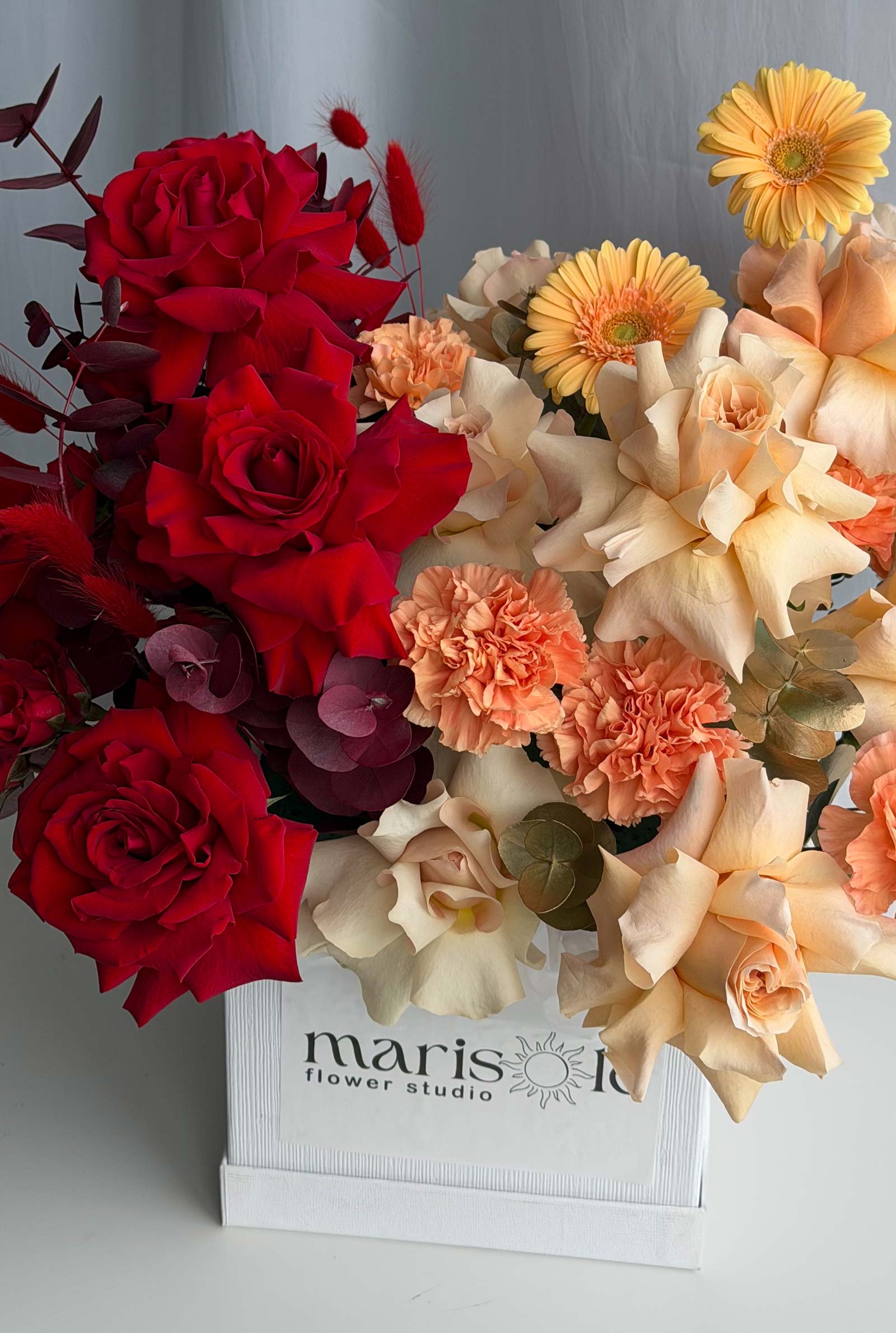 A vibrant floral arrangement featuring deep red roses, peach carnations, cream roses, and yellow gerbera daisies in a white wooden box labeled 'marisole flower studio'.