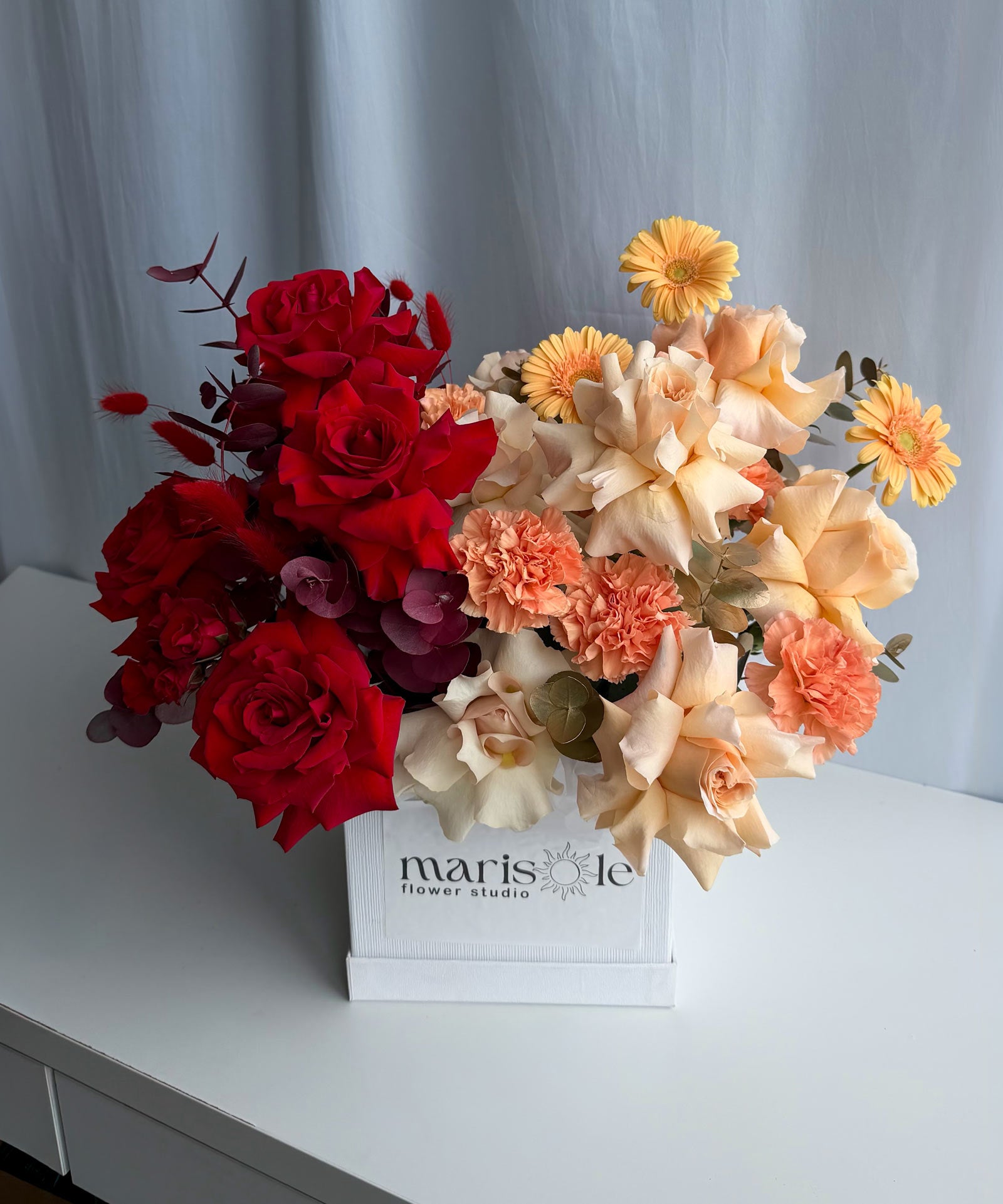 A floral arrangement in a white square box on a white table, featuring red roses, peach and cream flowers, and yellow daisy-like blooms against a light gray curtain background.
