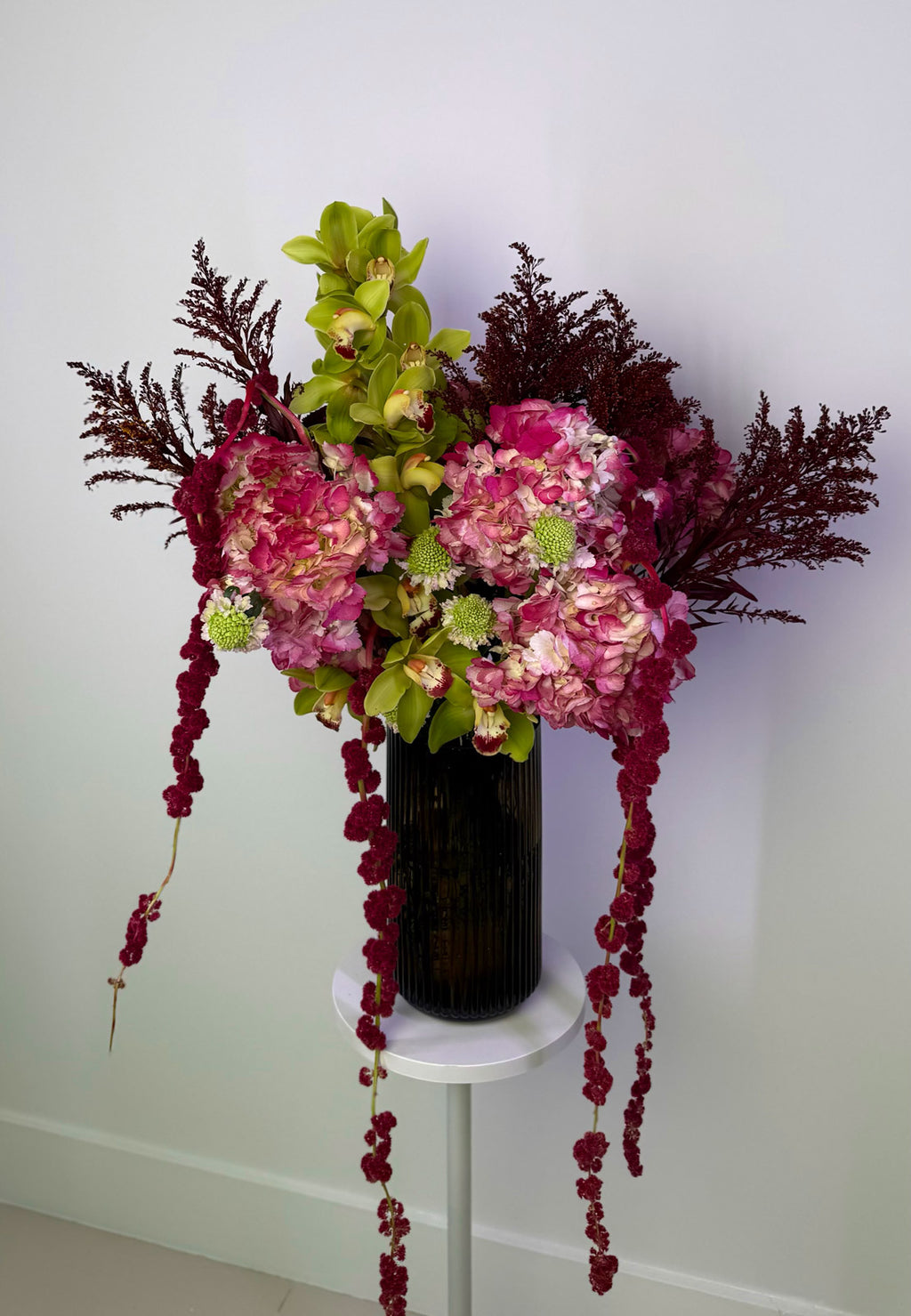 Floral arrangement with pink and green flowers in a black vase on a white pedestal against a white wall.