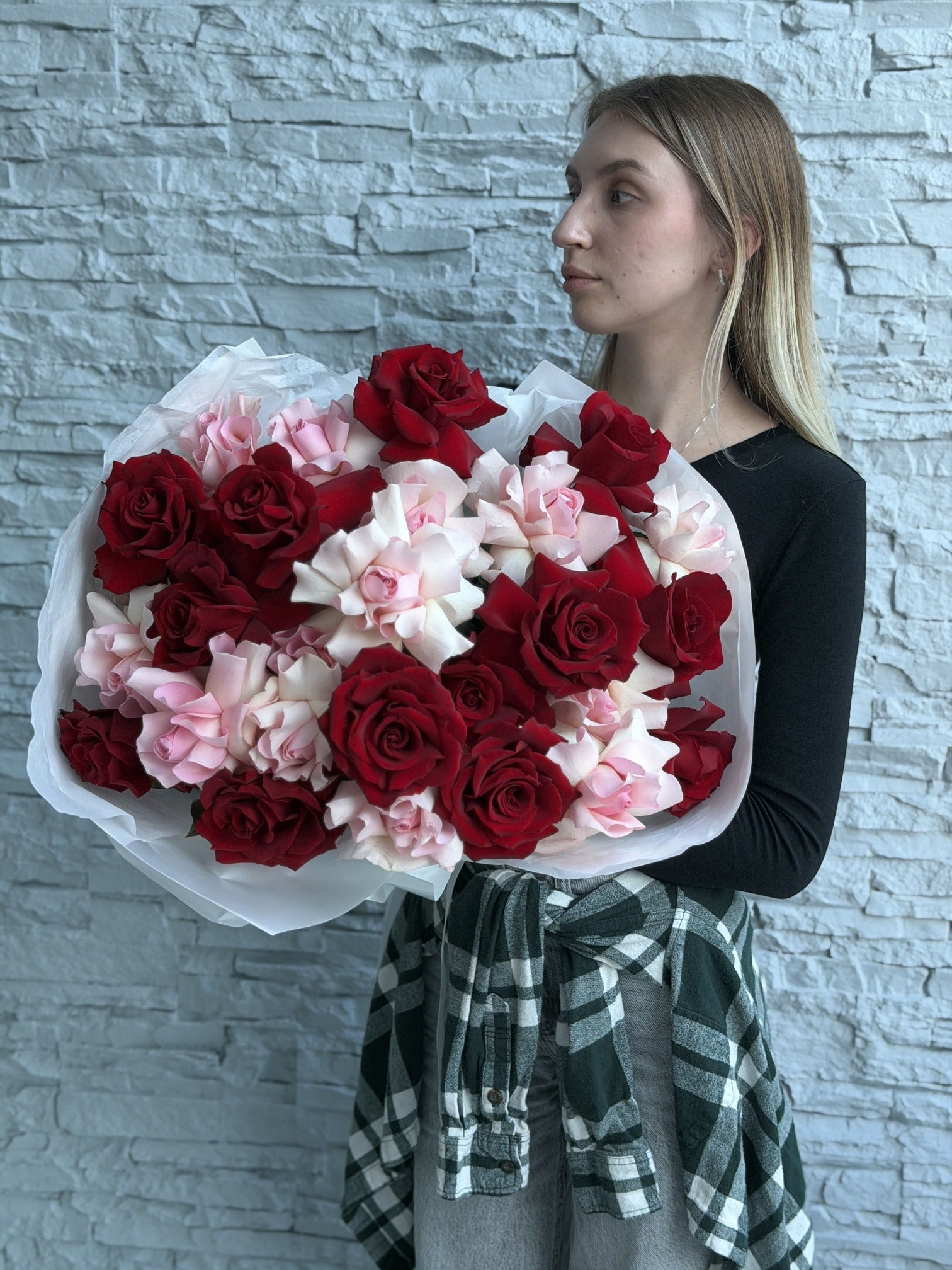 Woman holding a large bouquet of red and pink french roses against a stone wall.