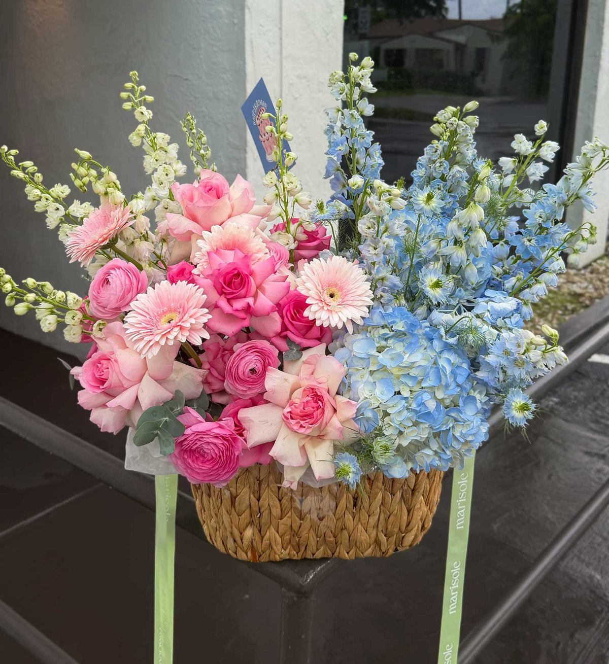 A woven basket floral arrangement featuring pink roses, pink ranunculus, light pink gerbera daisies, and light blue delphiniums, placed on a dark ledge in front of a building.
