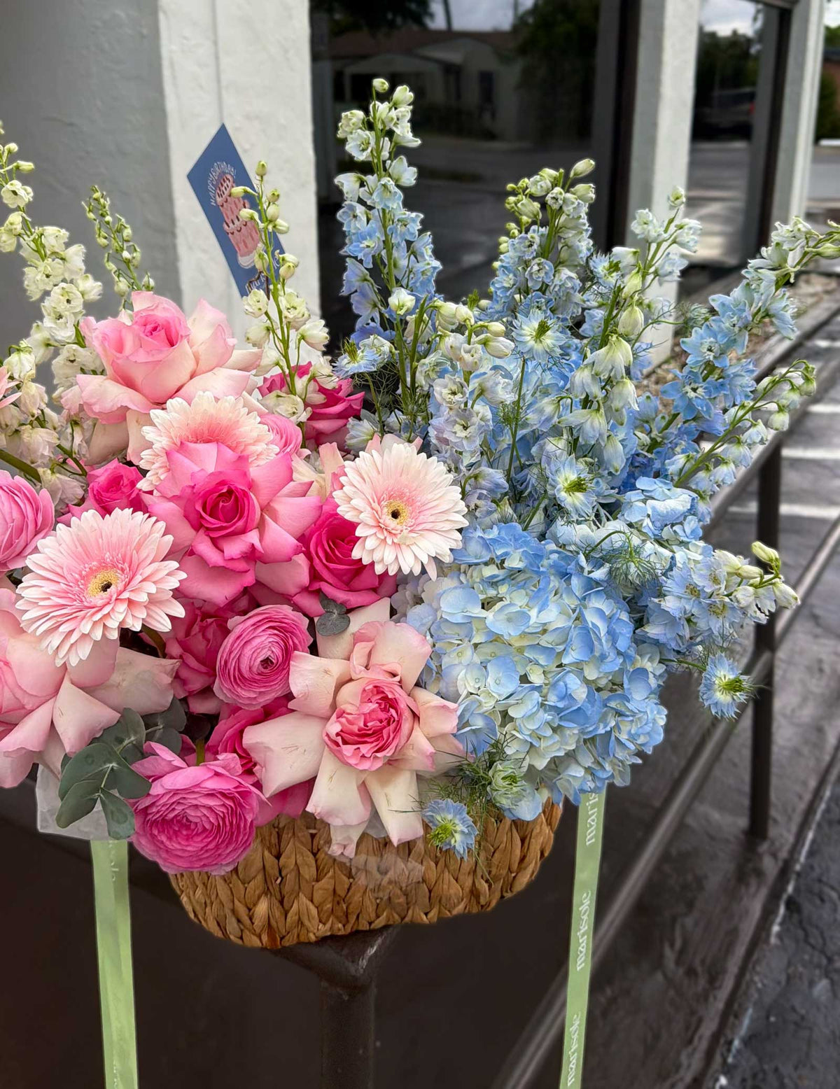 A woven basket filled with a mixed floral arrangement featuring pink roses, pink gerbera daisies, light pink ranunculus, and light blue delphiniums, displayed on a storefront window ledge.