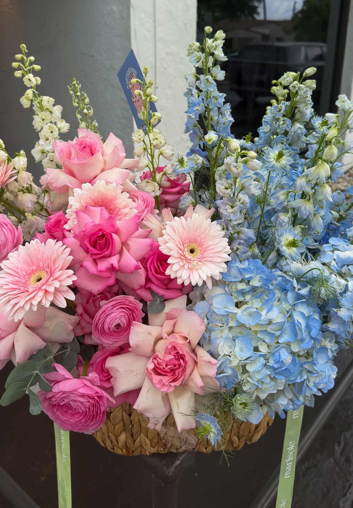A vibrant floral arrangement in a woven basket, featuring pink roses, pink and white gerbera daisies, light blue hydrangeas, and tall light blue delphiniums, set against a dark window background.