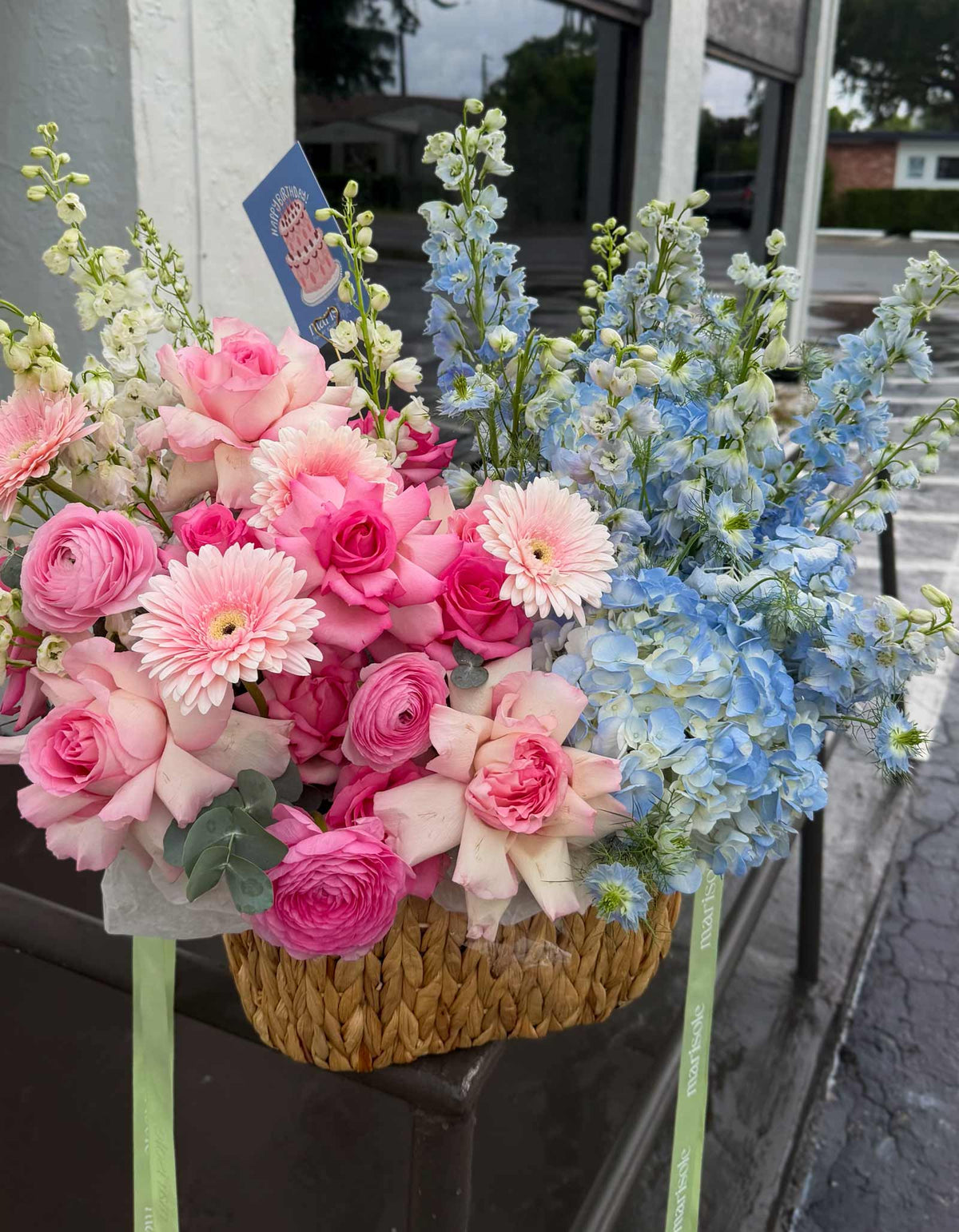 A vibrant floral arrangement in a woven basket, featuring pink and light pink roses, pink and white gerbera daisies, and light blue delphiniums, set on a storefront window ledge.
