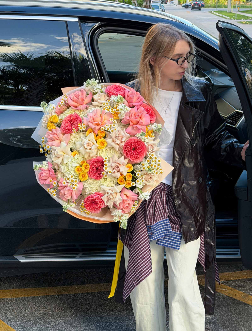 Woman standing beside an open car door holding the Golden Hour Bouquet with pink roses, ivory hydrangeas, yellow spray roses, and chamomile.