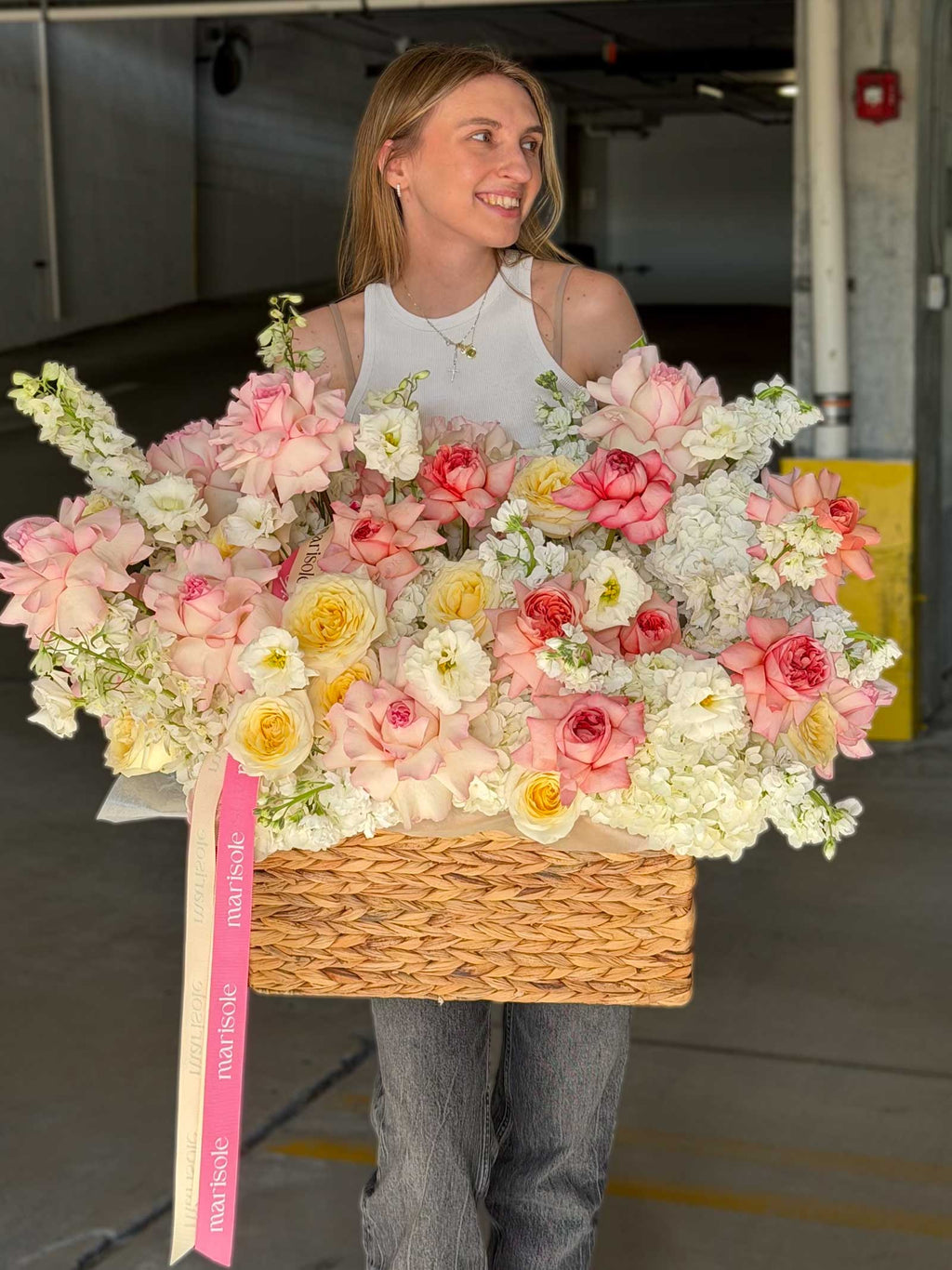 A woman in a white sleeveless top and gray jeans holds a large woven basket filled with pink, peach, cream, and yellow flowers. A pink ribbon with the brand name 'Flowers Direct' hangs from the side.