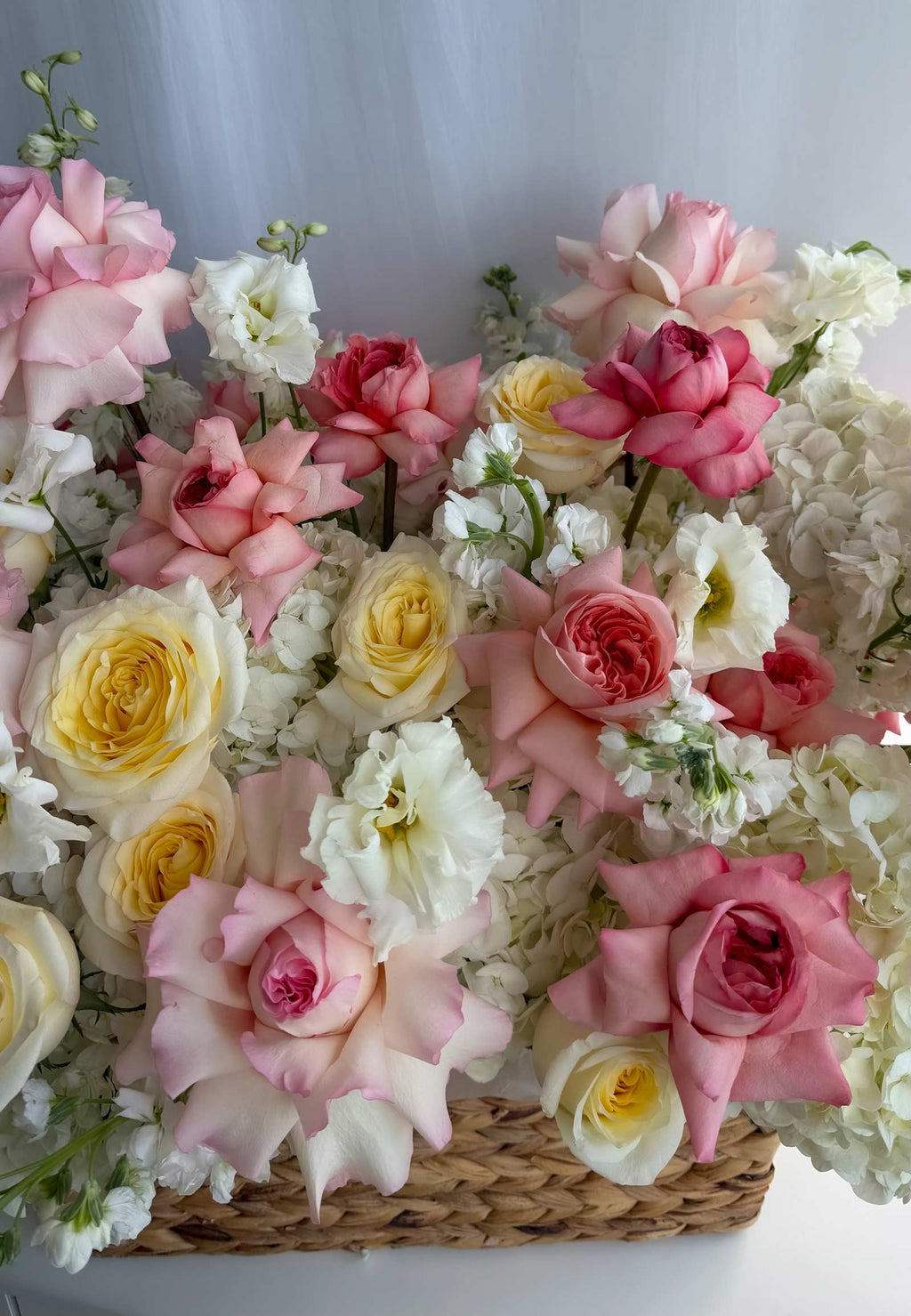 A dense floral arrangement of pink, white, and yellow roses, hydrangeas, and small white flowers in a woven basket, photographed against a soft light gray background.